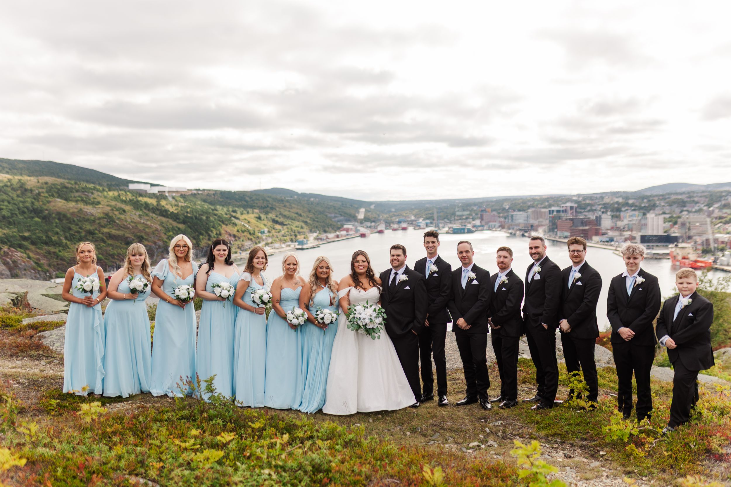 Wedding party members pose together against a scenic mountain and water backdrop in pastel blue and black attire.