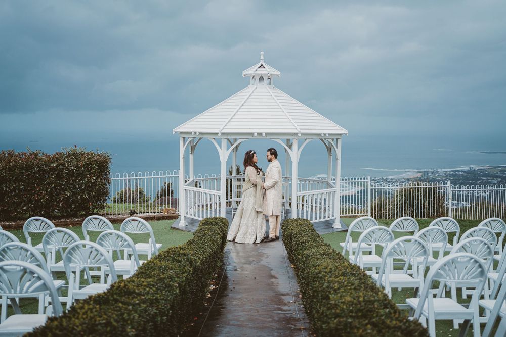 Elegant white wedding gazebo and chairs set up on a grassy hilltop with ocean views in background.