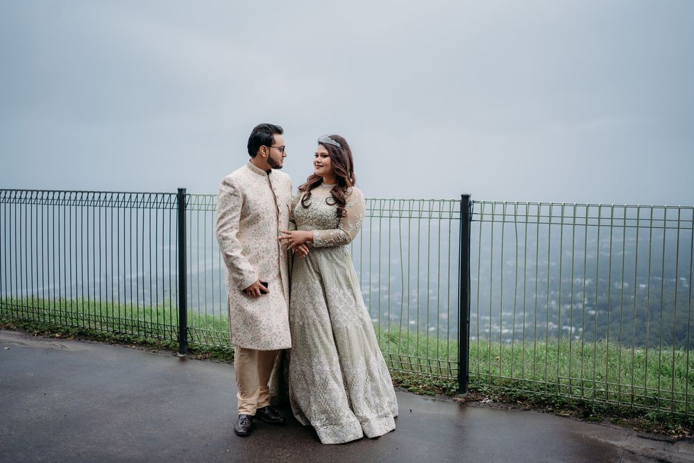 Couple in light-colored outfits standing on a scenic overlook with metal railing and foggy views in the background.