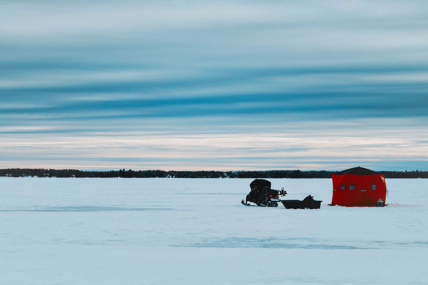 A red ice fishing shelter and snowmobile sit on a frozen lake under a soft blue winter sky.