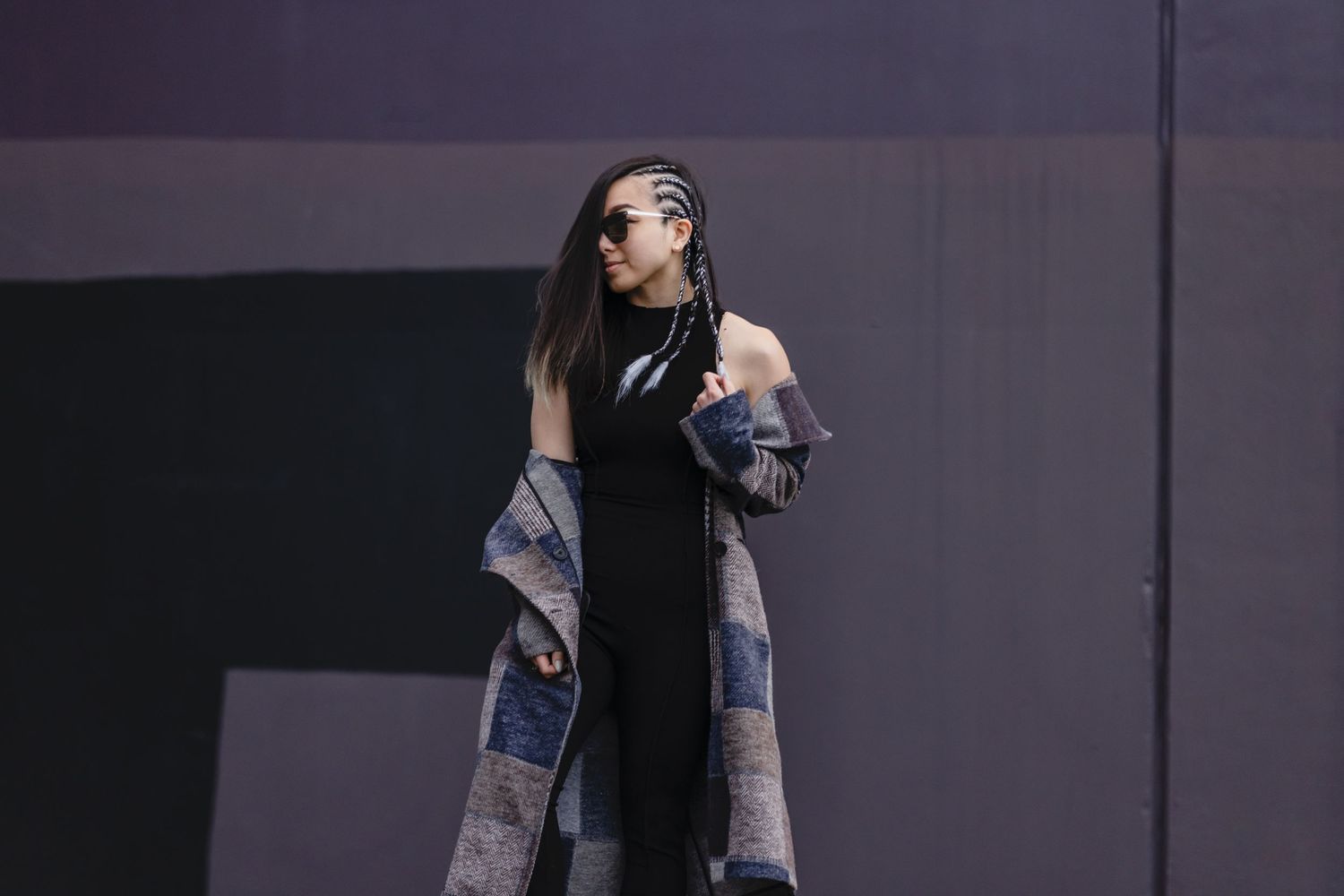 A person in a black dress and patterned long cardigan poses against a dark wall in dramatic lighting.
