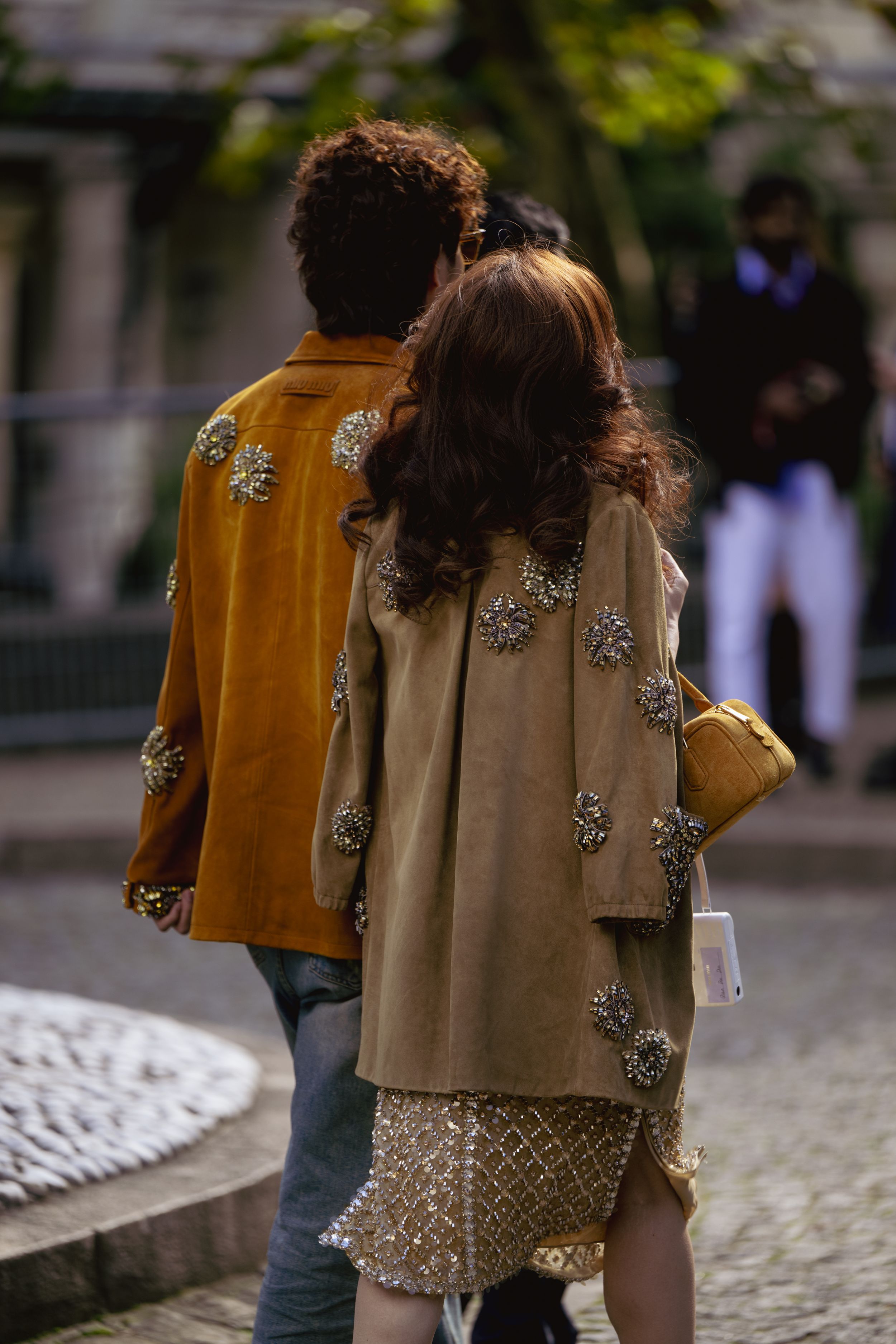Couple wearing embellished coats in mustard and beige walk together on cobblestone street.