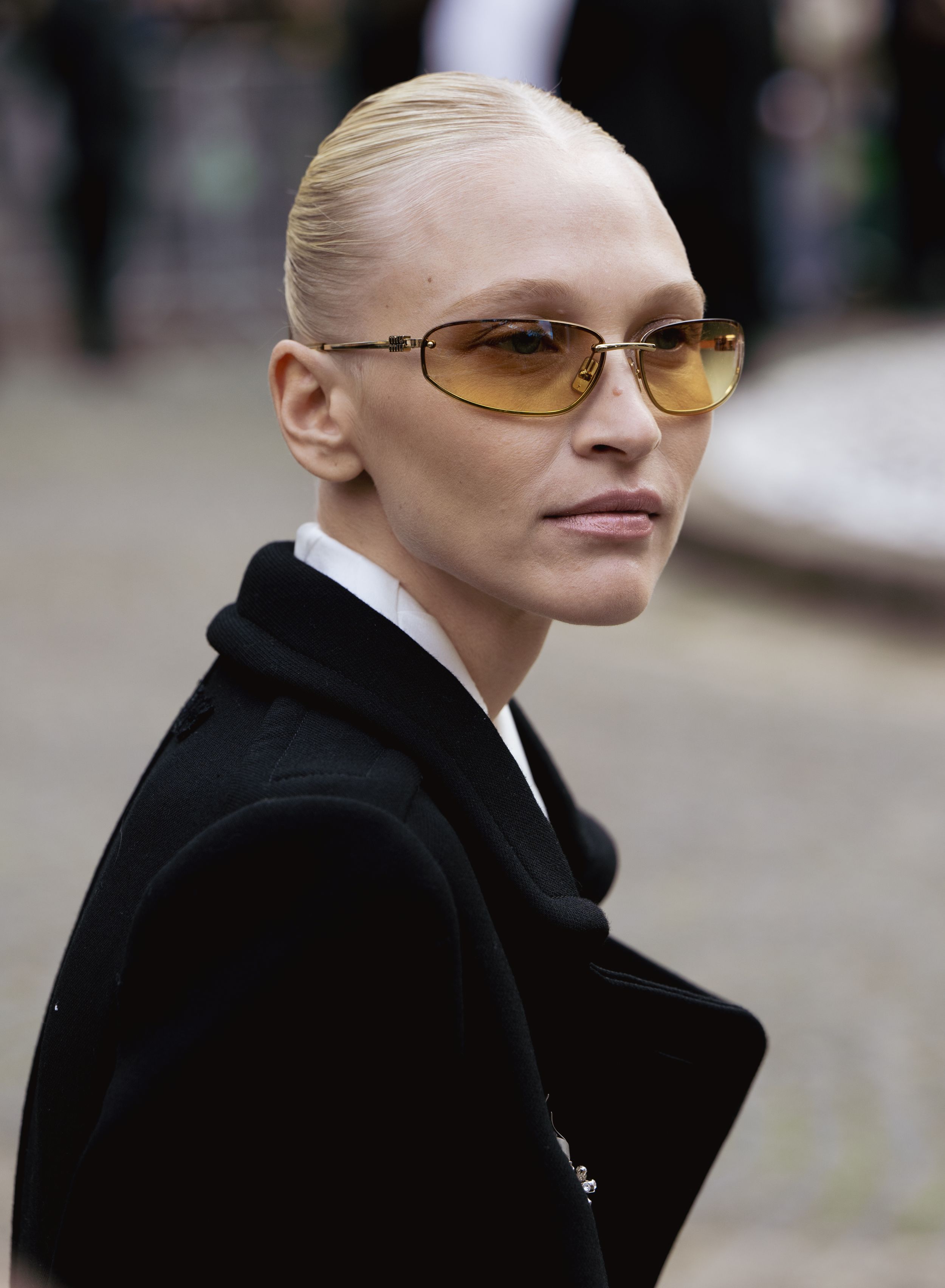 Person wearing amber-tinted glasses and black coat poses against blurred urban background.