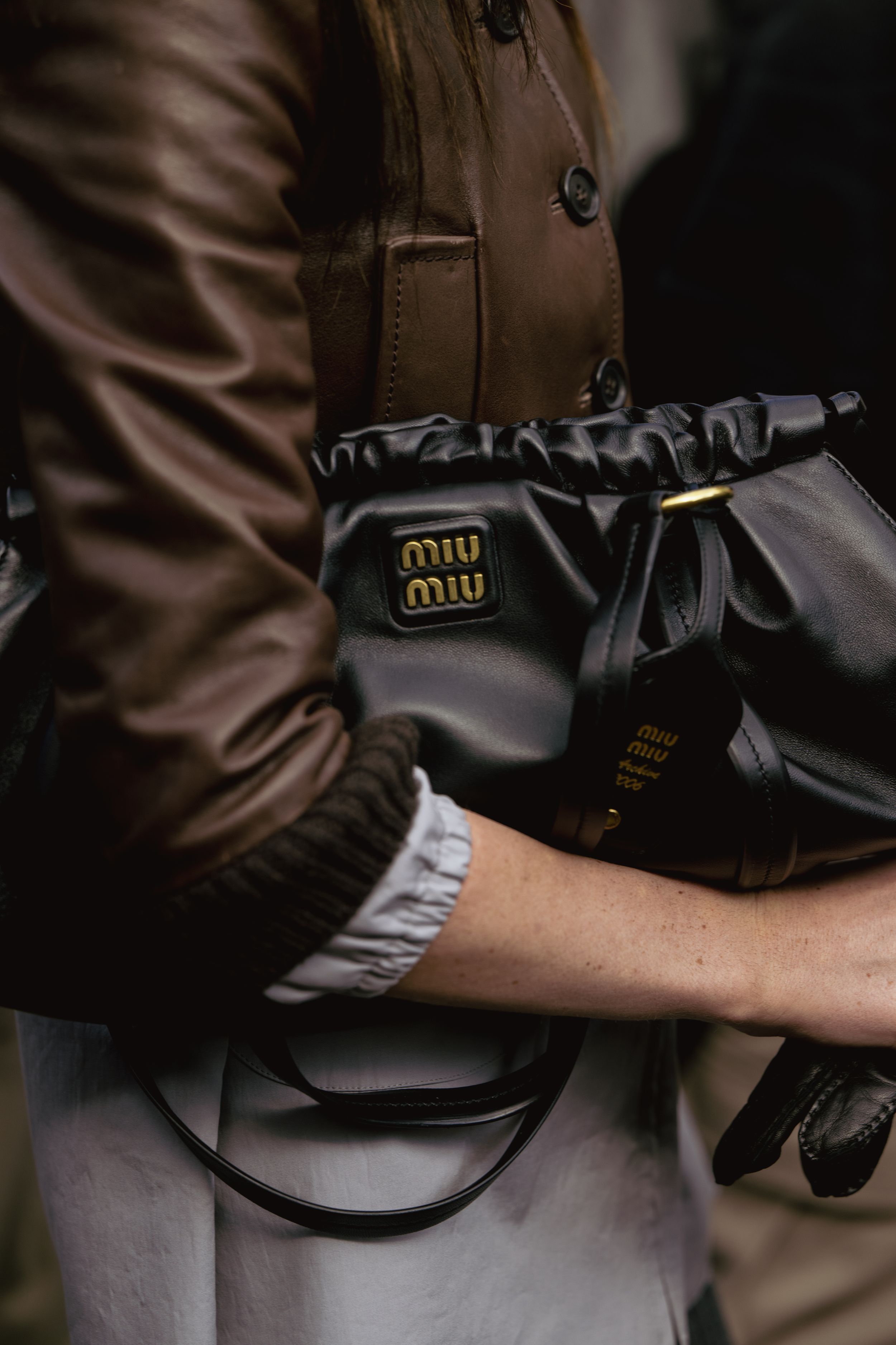 A close-up of a black Miu Miu handbag being held against a brown leather jacket.