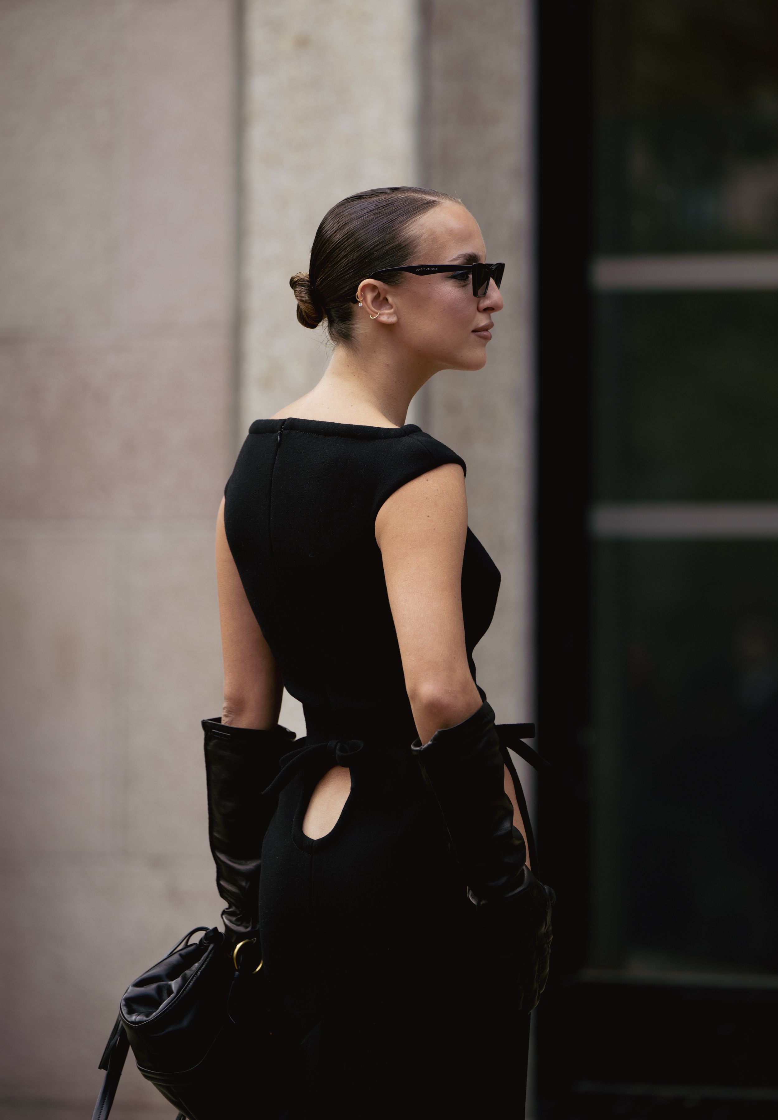 Elegant minimal black dress with cutout details and gloves seen from behind on a city street.