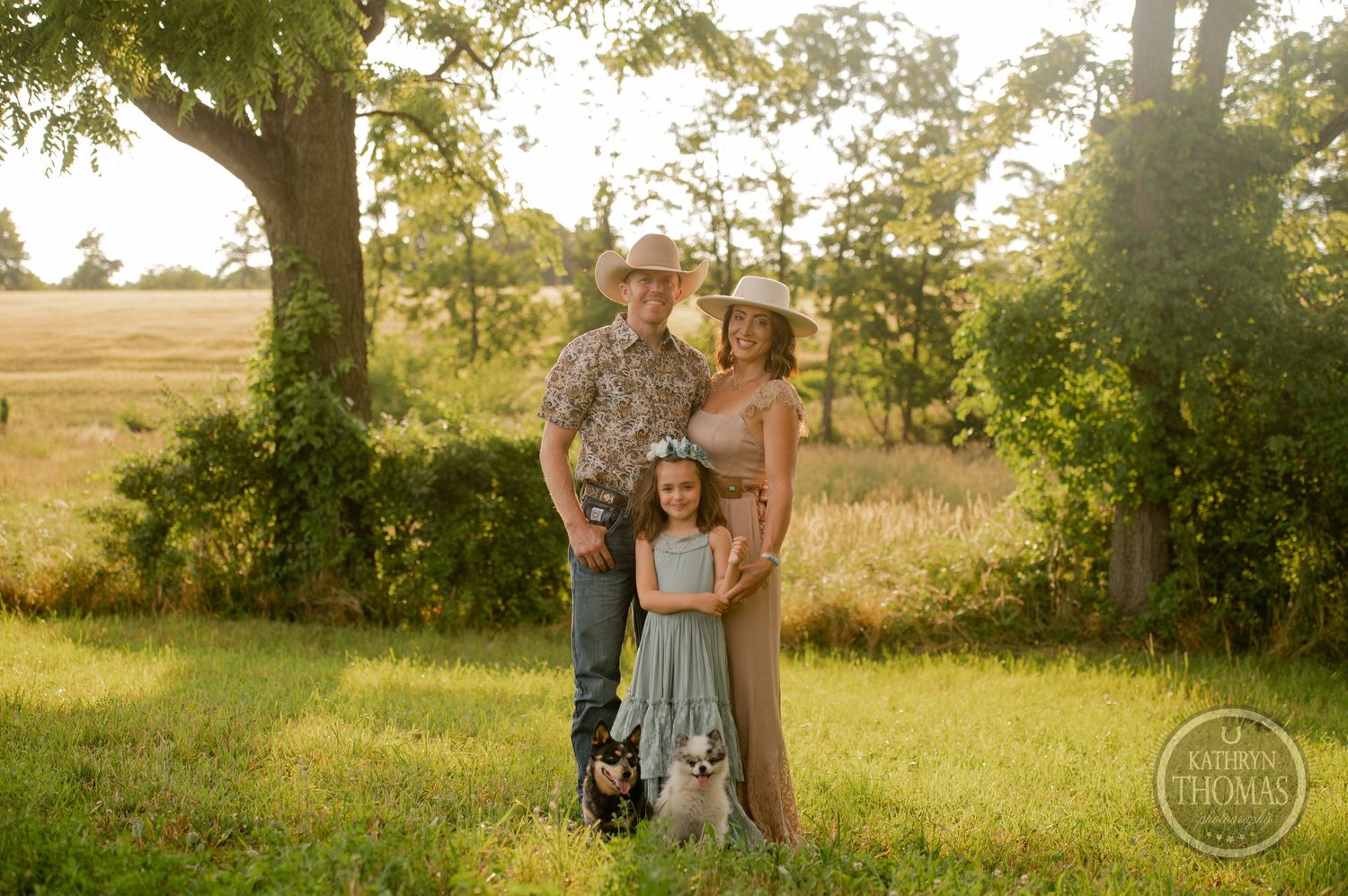 Family portrait in golden evening light with western attire and two small dogs in a rural setting.