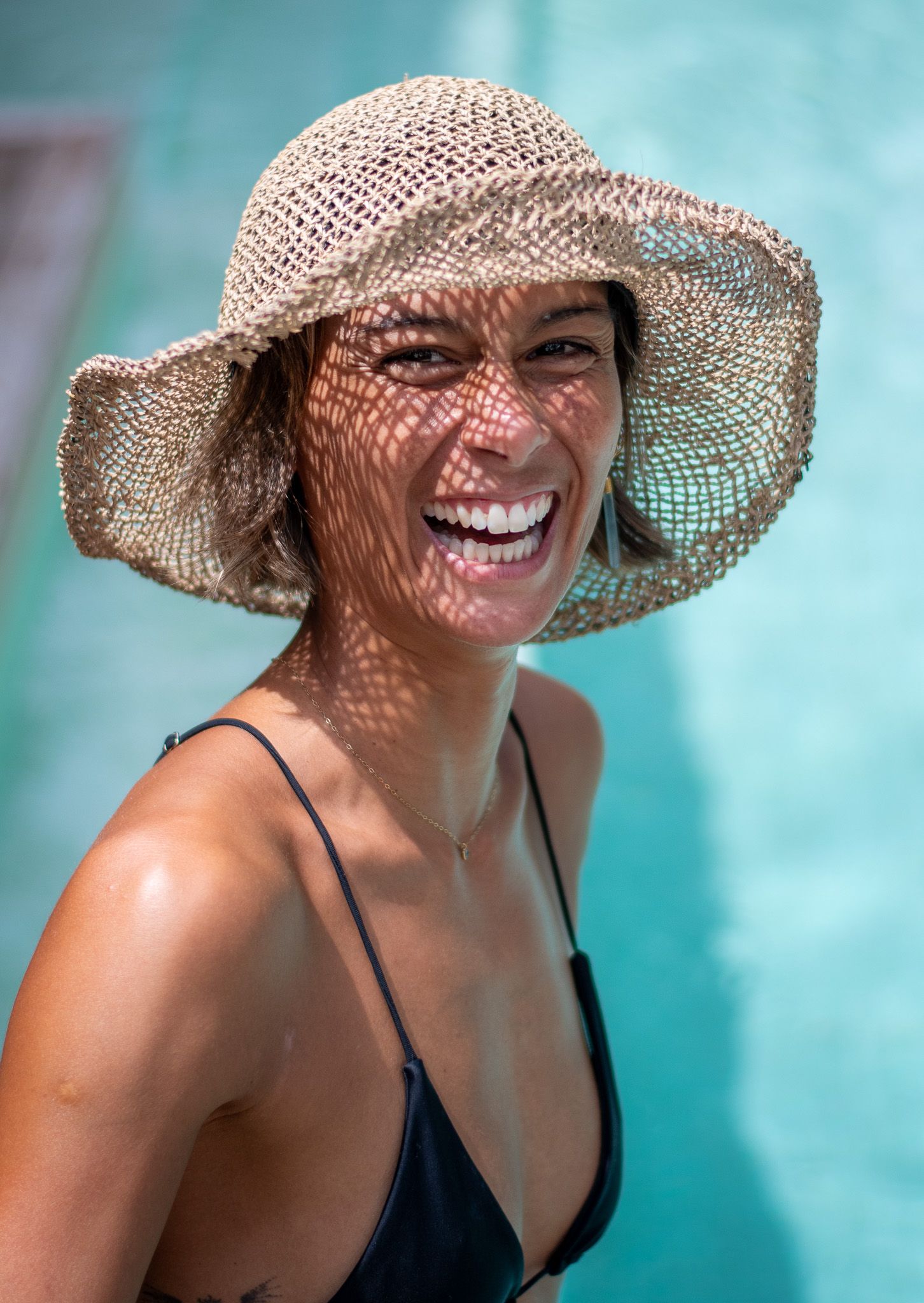 A joyful person in a wide-brimmed straw hat and black swimsuit smiles against a turquoise background.