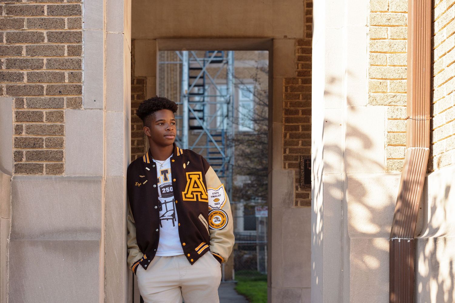 A person wearing a varsity jacket leans against brick pillars in warm sunlight.