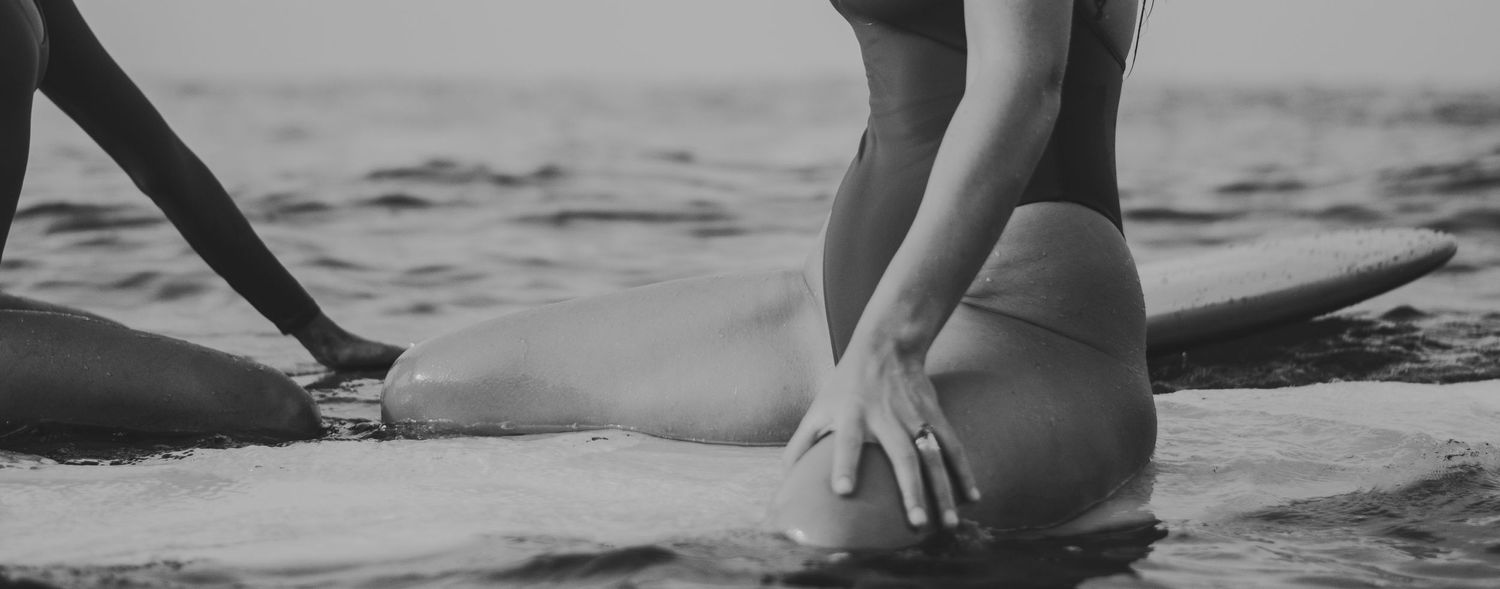 Black and white photo of a surfer touching the sand while sitting on a beach with ocean waves in the background.