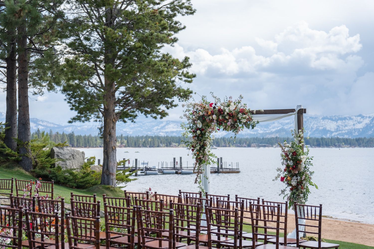 Wooden wedding ceremony setup at Edgewood with floral arch and chiavari chairs on a Lake Tahoe beach with mountains in the background.