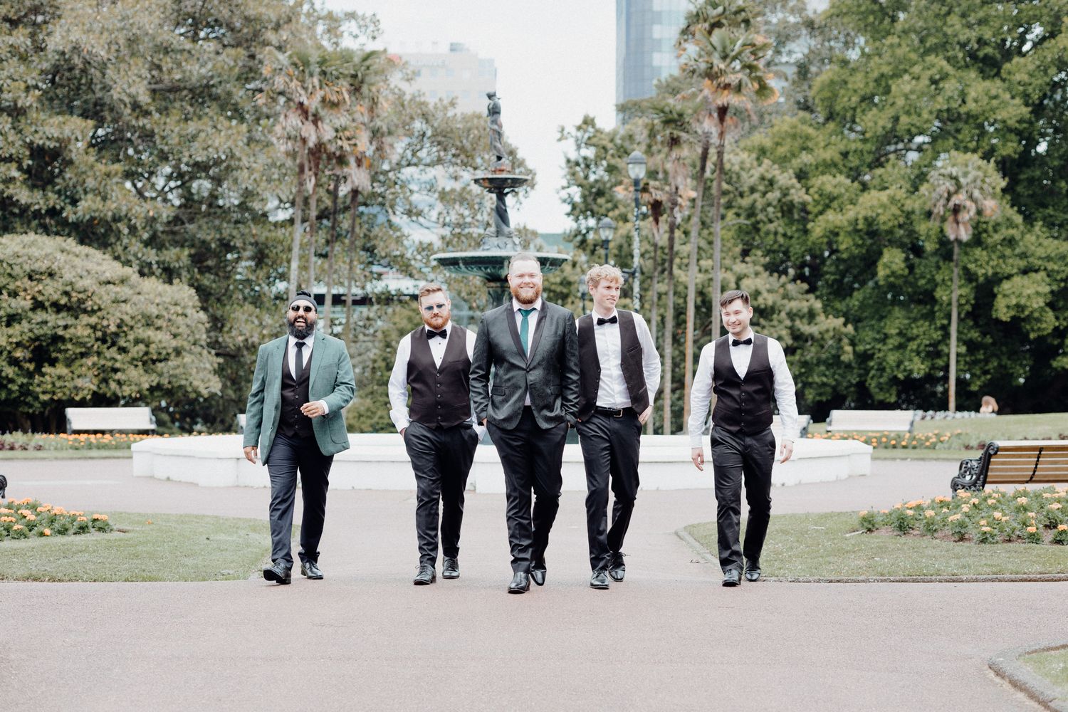 The groom's squad walk along a park pathway on a sunny day, captured by Apertured Media Auckland Wedding Photographer.