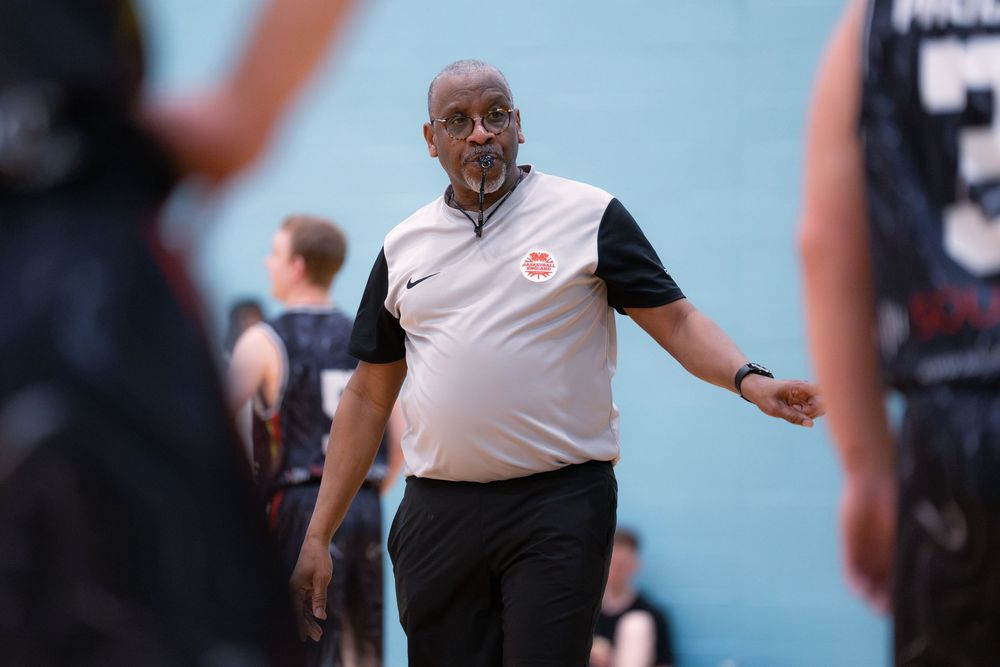 Coach in black and white polo shirt gesturing during indoor basketball tournament.