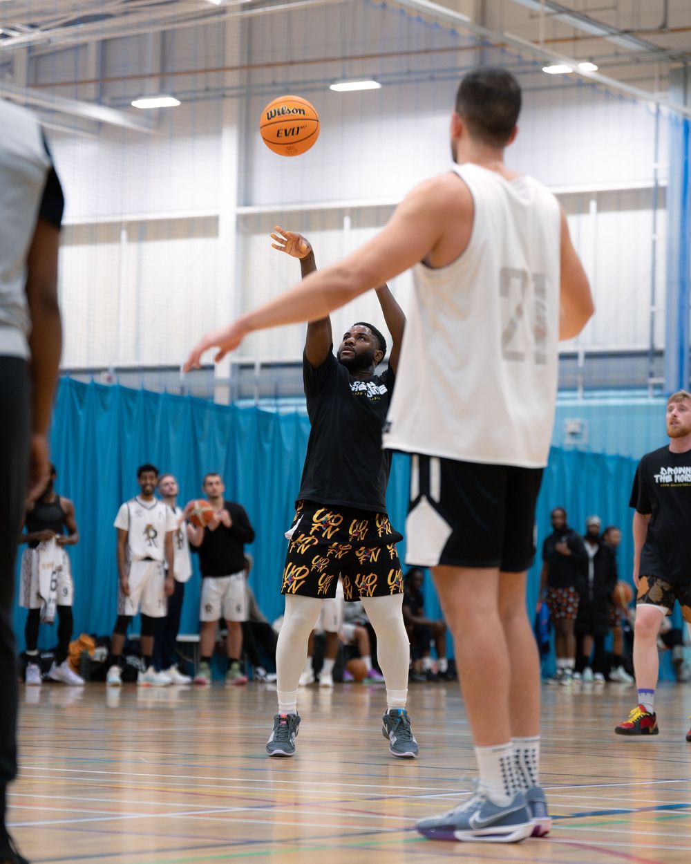 Basketball players practicing shooting drills in an indoor gym with blue curtains and a basketball flying overhead.