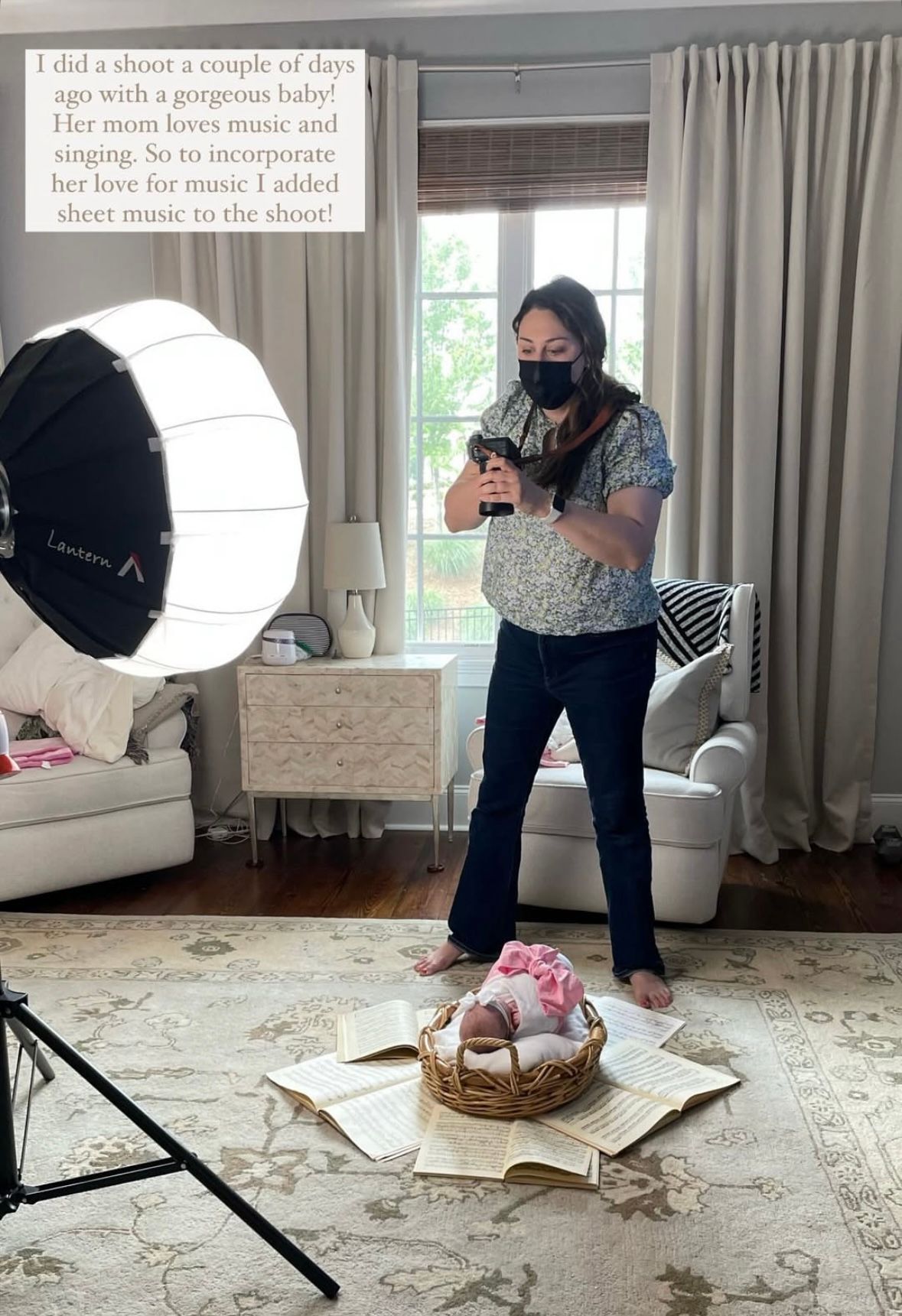 Newborn photographer in a mask photographing a baby in a basket surrounded by sheet music on a rug.
