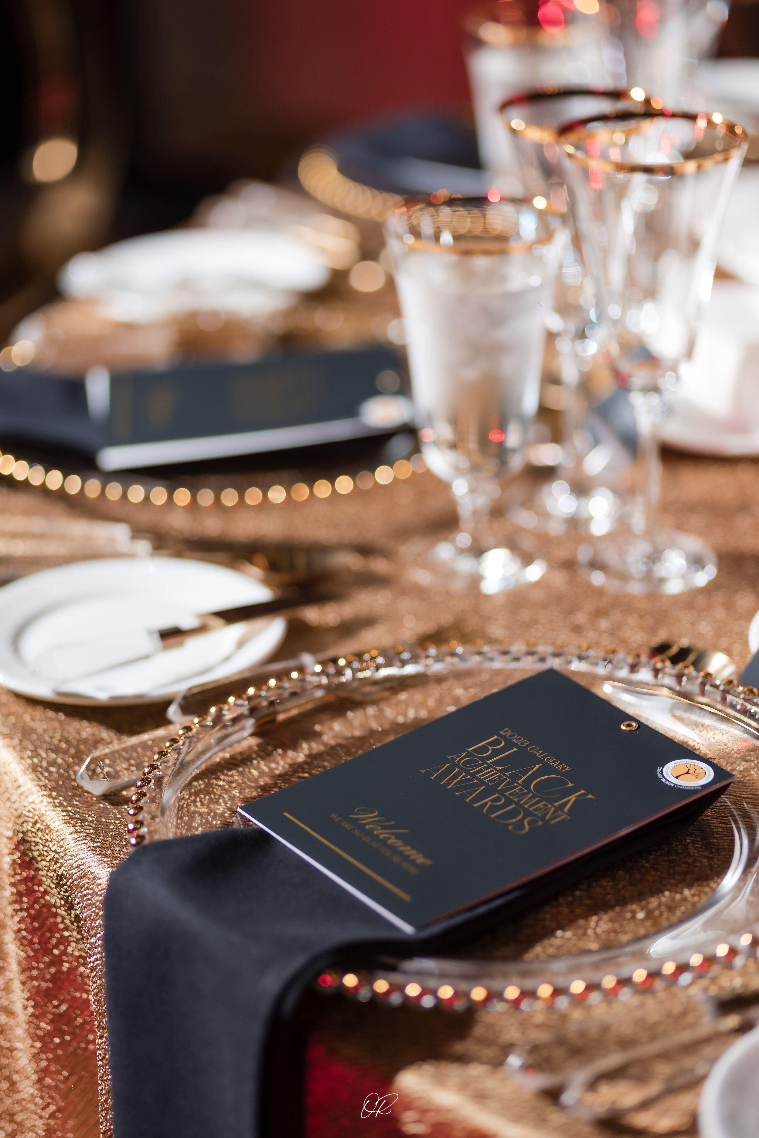 Elegant table setting with gold charger plates, black napkins and crystal glasses on a shimmering tablecloth.