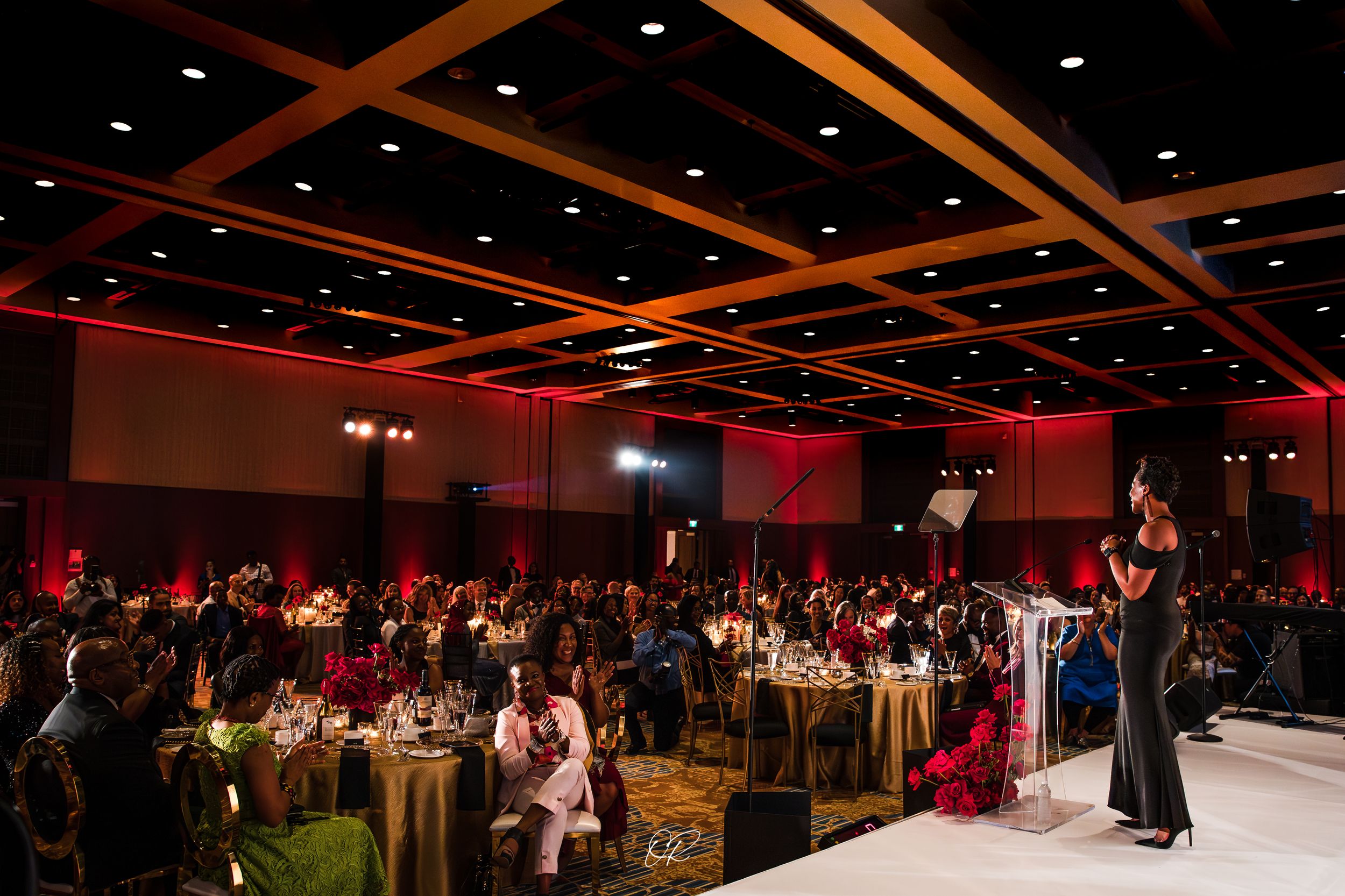 An elegant banquet hall with red ambient lighting features round dining tables and a speaker podium on a white stage.