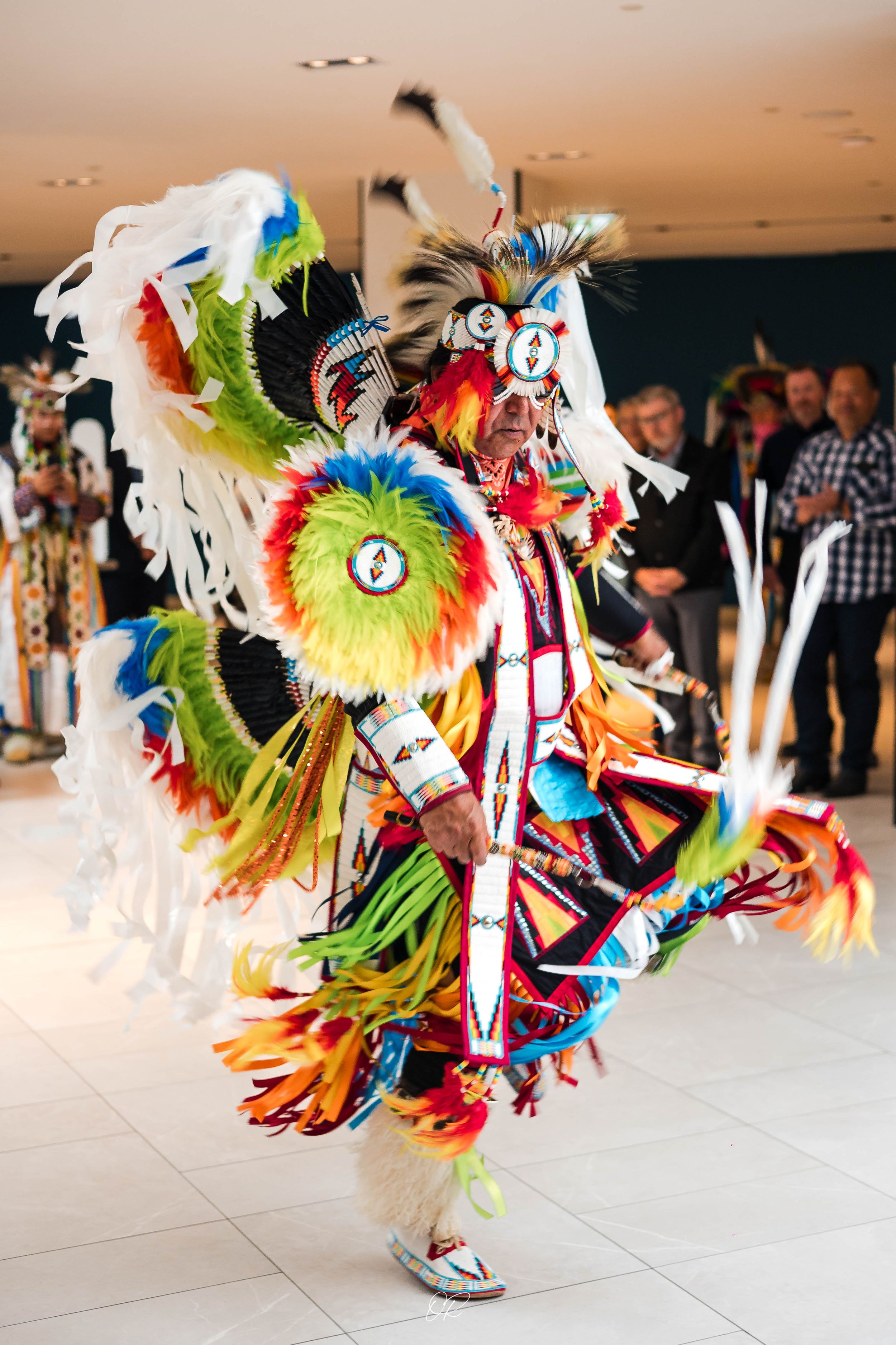 A dancer performs in vibrant traditional Native American regalia with colorful feathers and decorative garments at a powwow event.