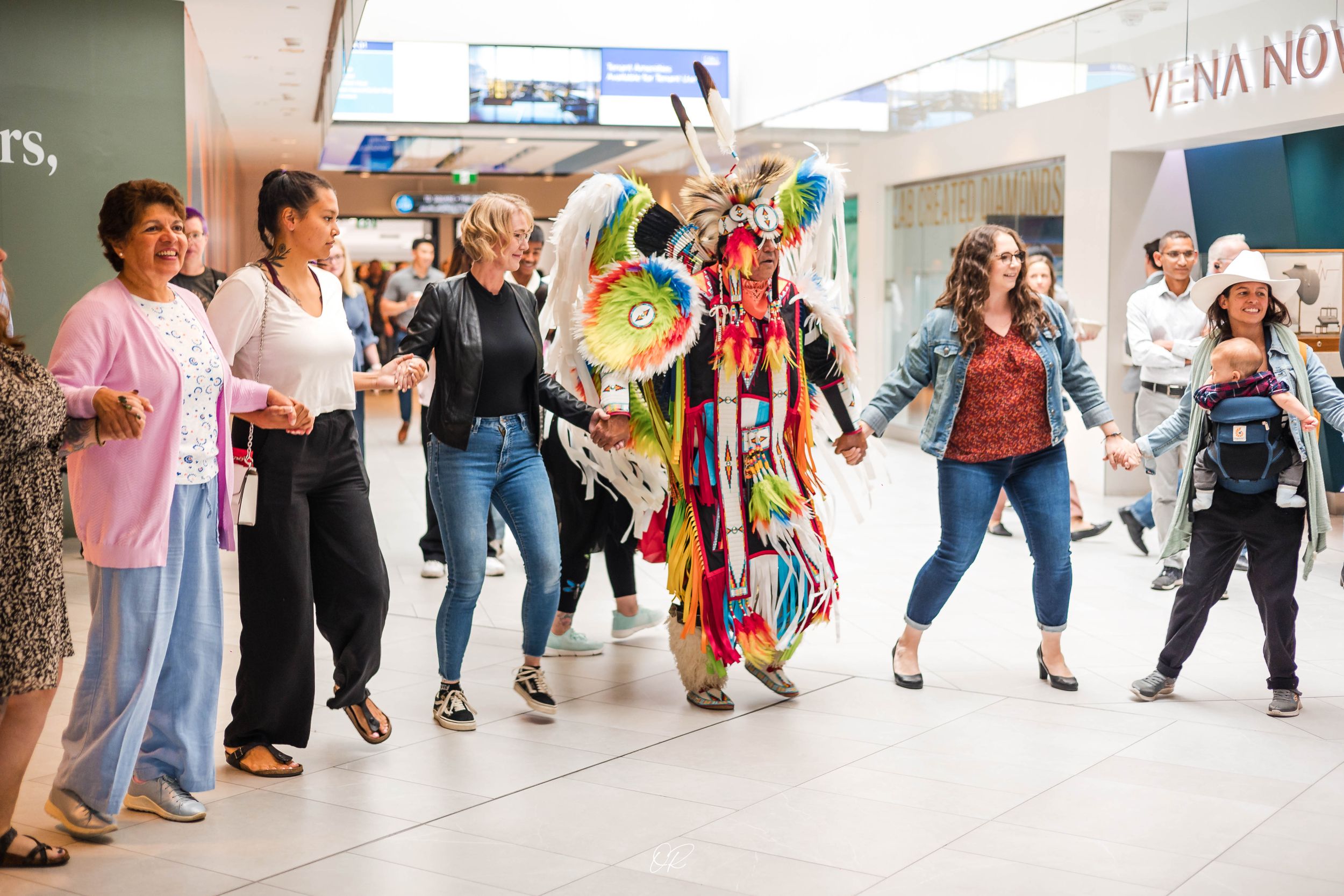 Colorful powwow dancer performs in traditional regalia at indoor shopping center while spectators watch.