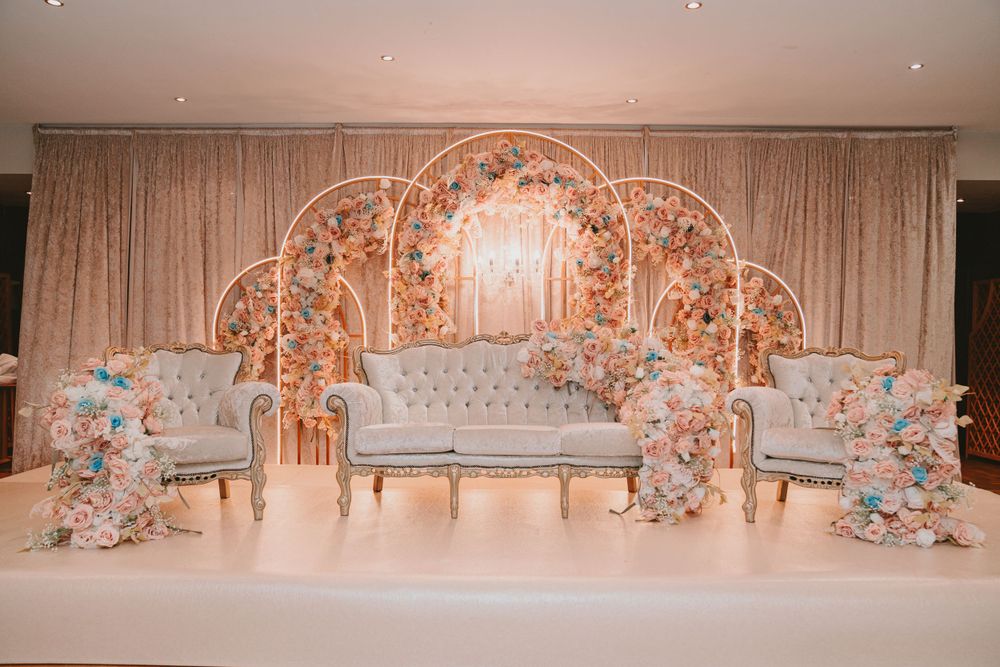 Elegant wedding stage setup with ornate gold arch backdrop, grey sofa seating and floral decorations in pastel colors.