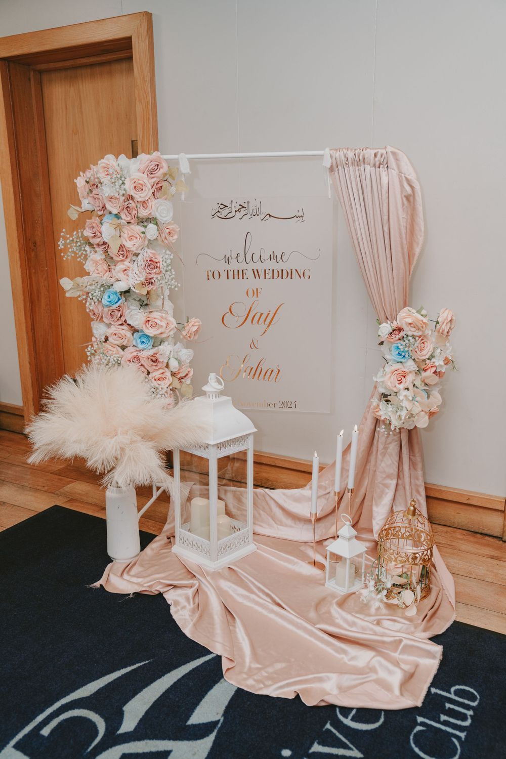 Wedding welcome sign with Arabic calligraphy adorned with blush and blue flowers, lanterns and draped fabric.