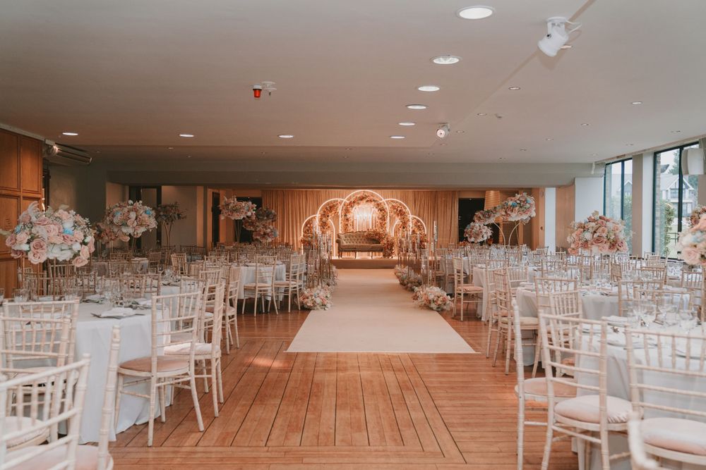 Long wedding ceremony aisle with white chairs and elegant gold arch backdrop in spacious venue.