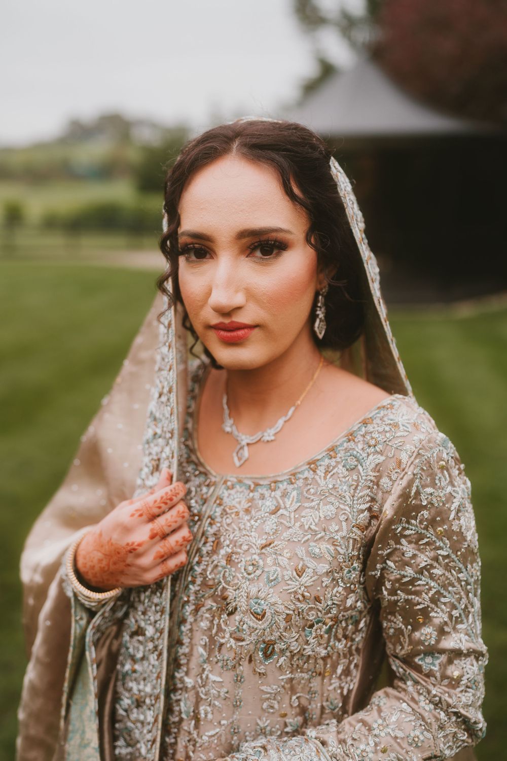 Bride in intricate beige lace gown poses with delicate veil outdoors.
