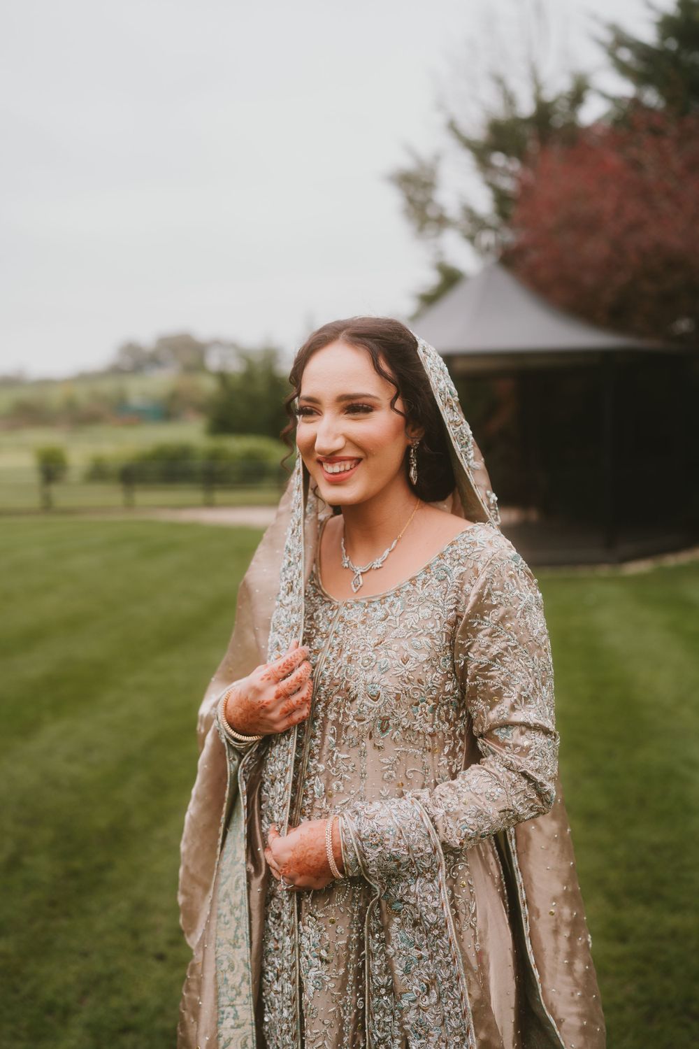 A bride wearing an ornate beige sequined dress and veil smiles while standing on manicured grass.