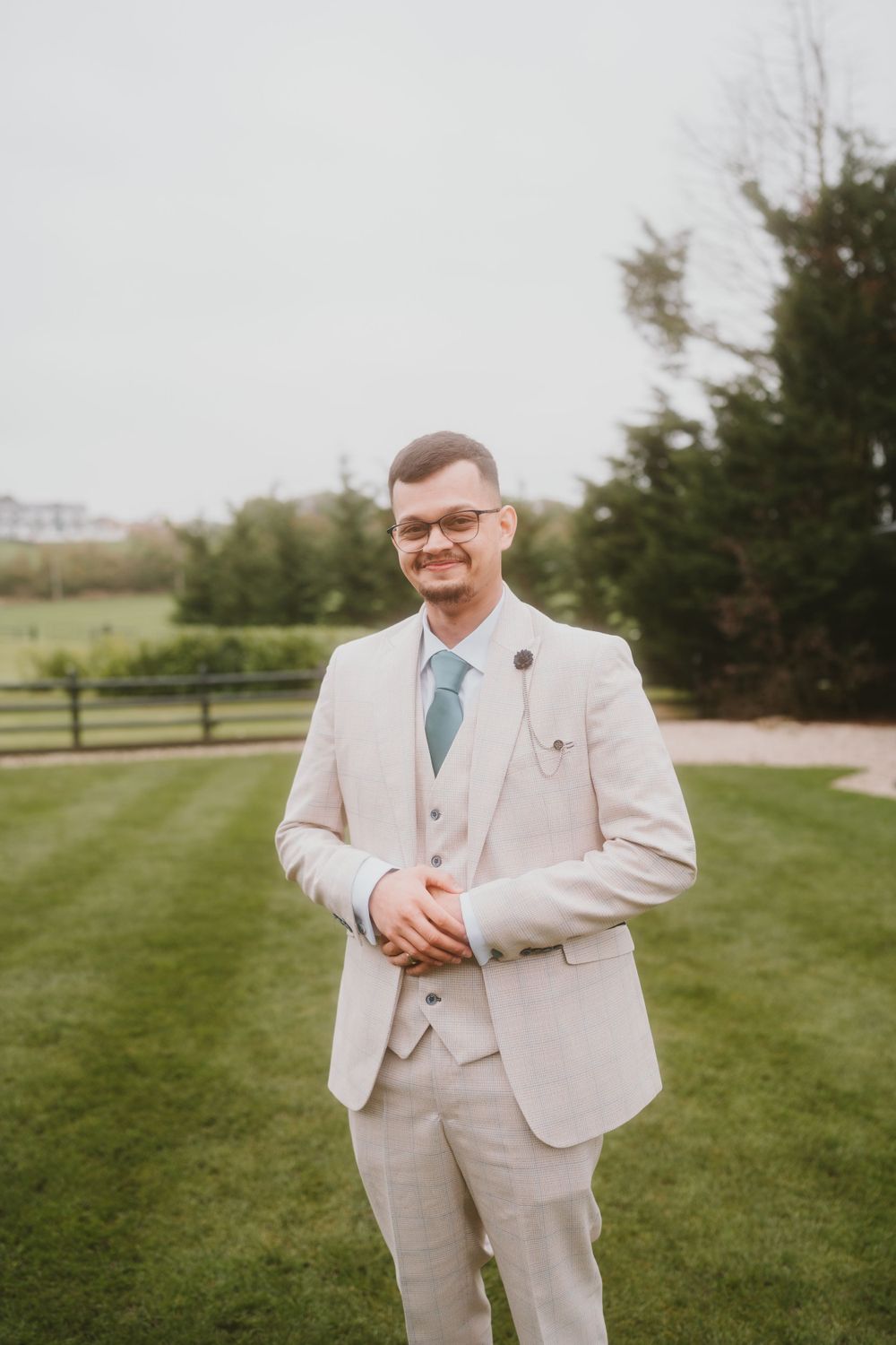 Groom in light beige suit with blue tie poses on lawn with autumn trees in background.