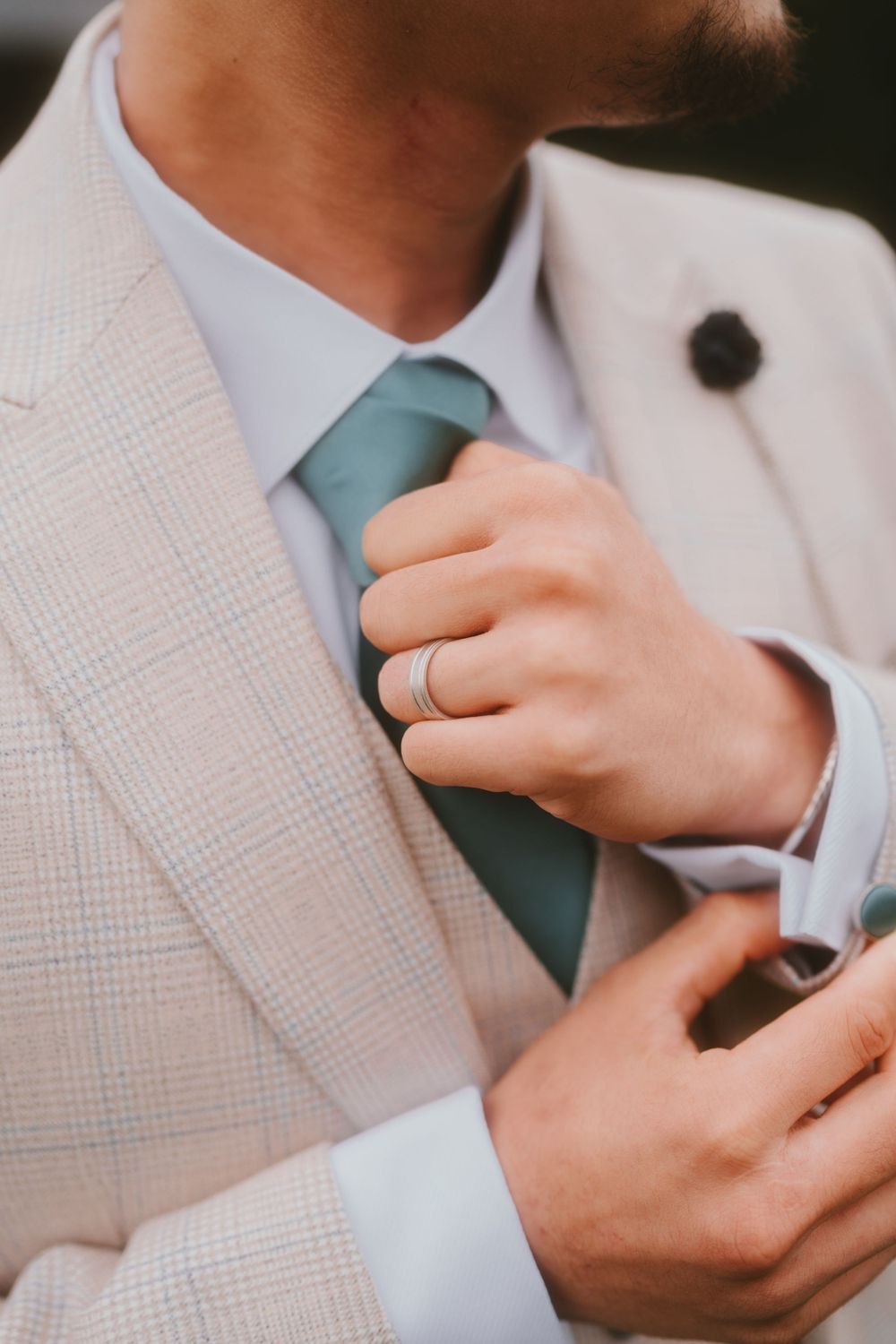Close-up of groom's hands adjusting blue tie, showing wedding ring and beige suit jacket.
