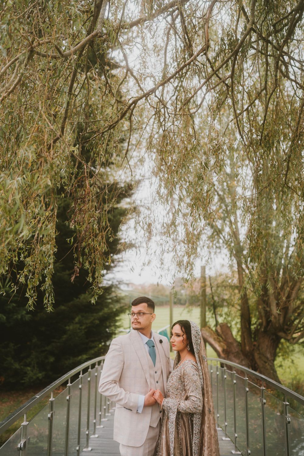Wedding couple posing on metal bridge with willow trees creating romantic backdrop.