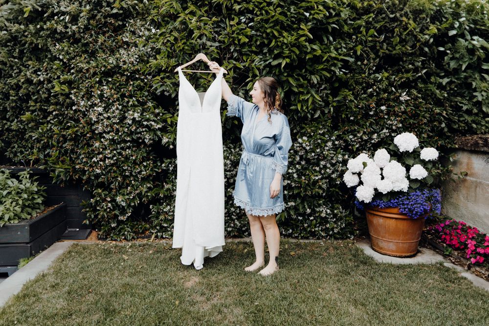 Bride holding her dress in a garden setting next to potted flower, captured by Apertured Media Auckland Wedding Photographer.