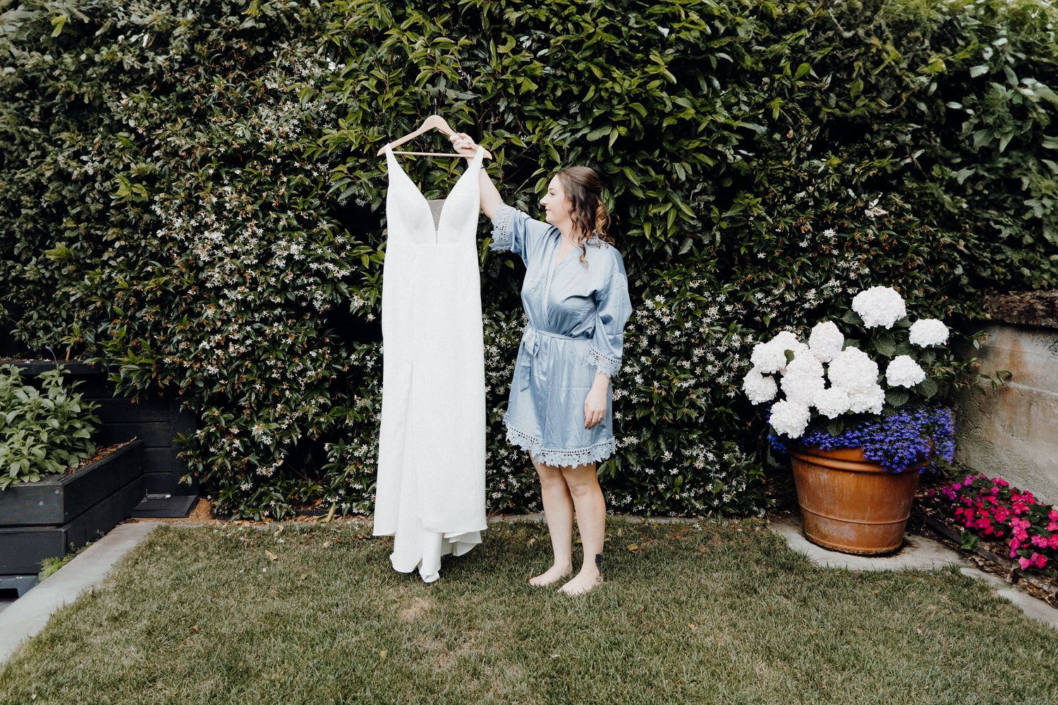 Bride holding her dress in a garden setting next to potted flower, captured by Apertured Media Auckland Wedding Photographer.