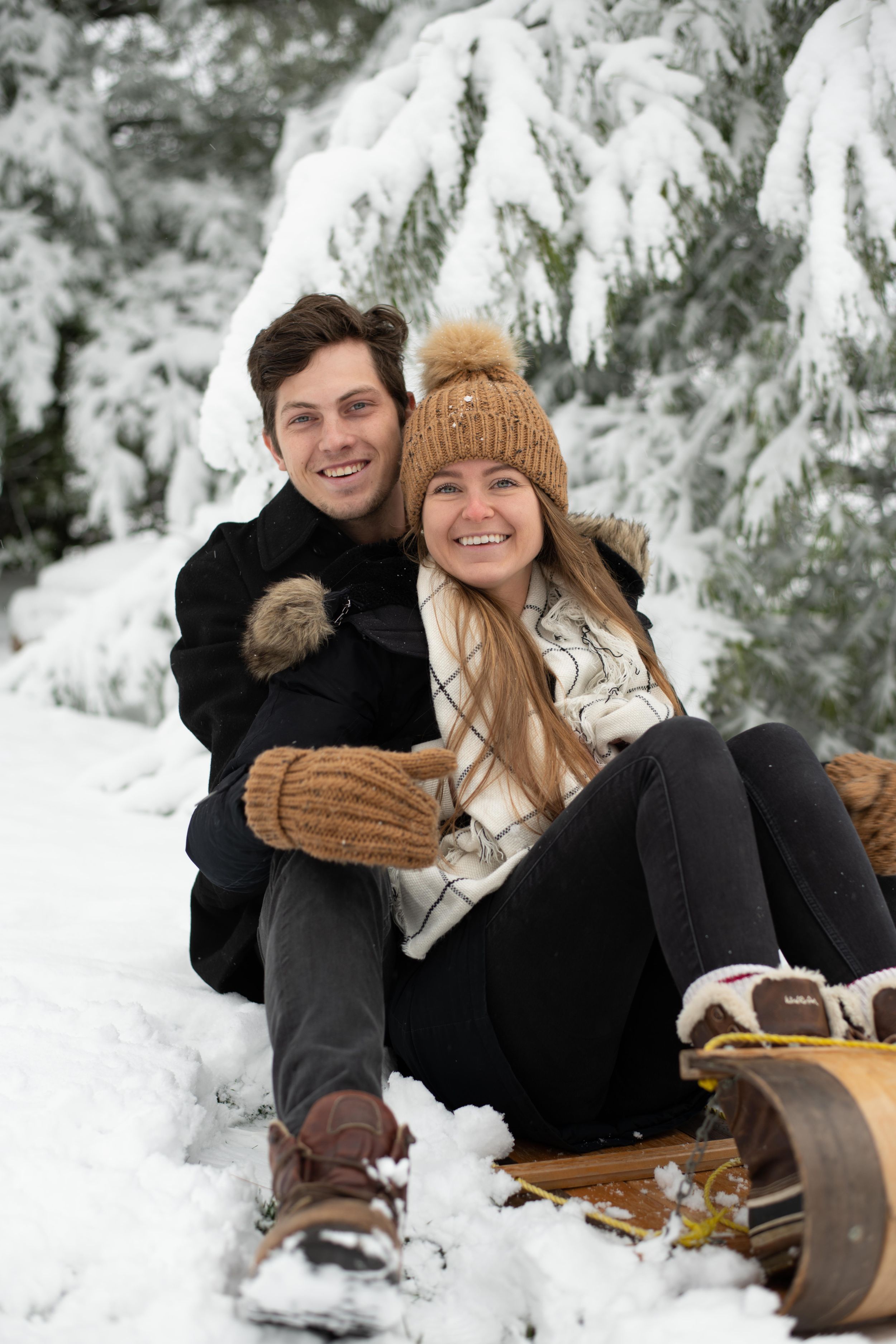 A couple sitting on a sled on a snow-covered ground, surrounded by snow-laden evergreen branches.