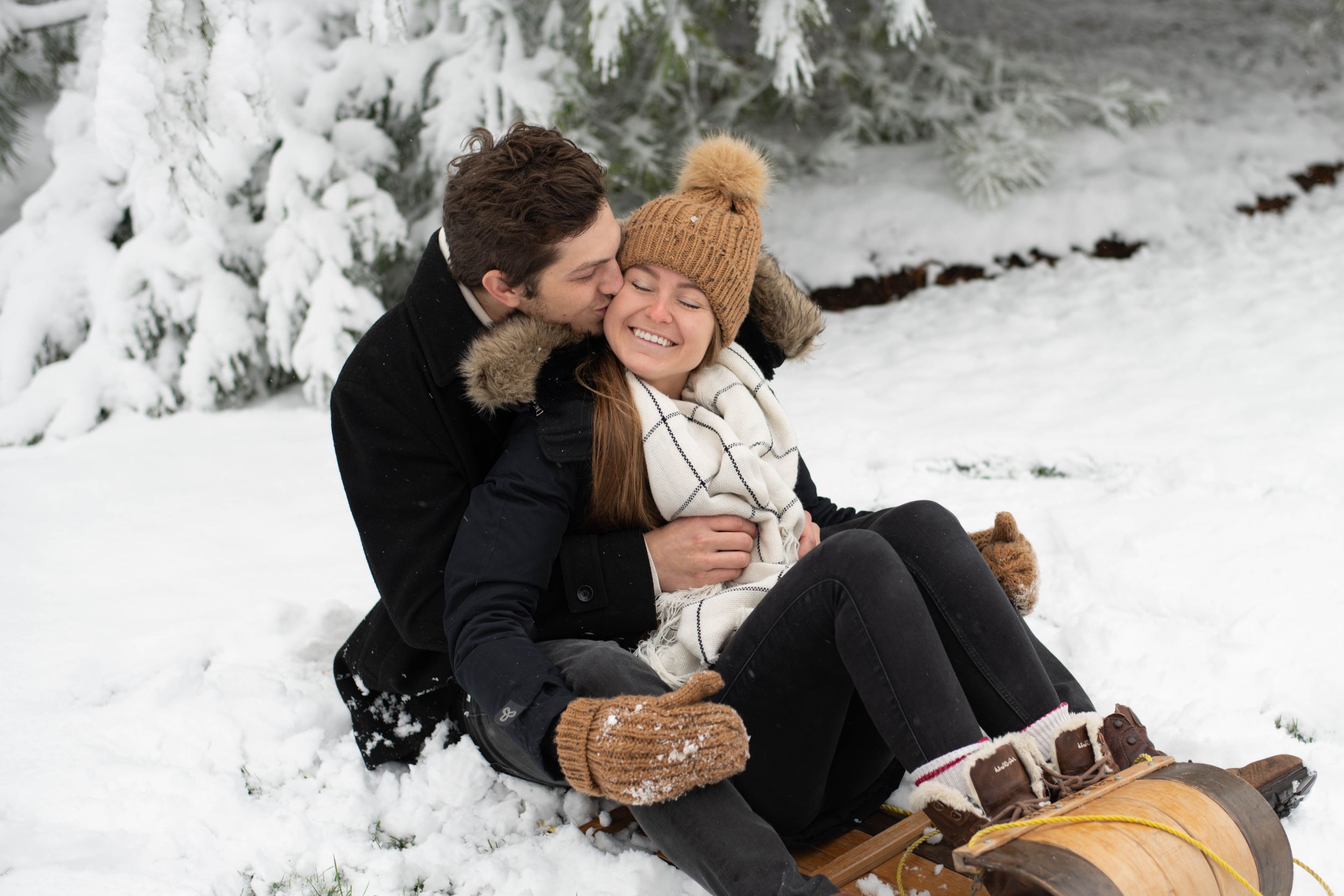 Romantic winter scene of couple embracing in snow near snow-covered evergreen trees sitting on a wooden sled.