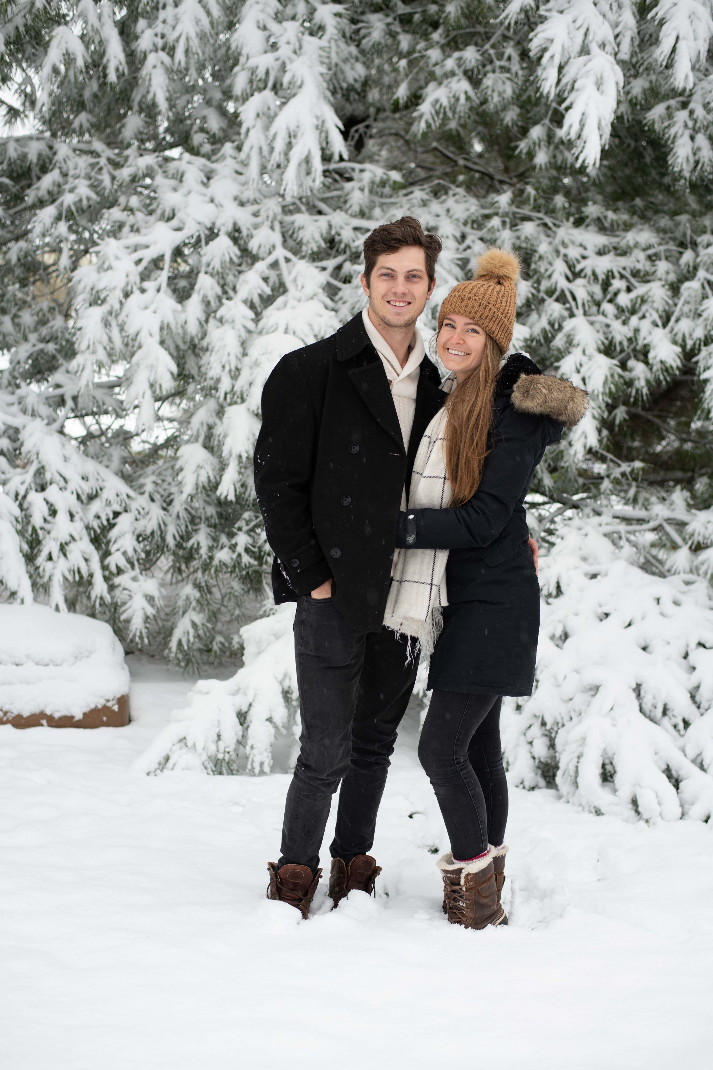 Two people in winter attire standing together against snow-covered evergreen trees.
