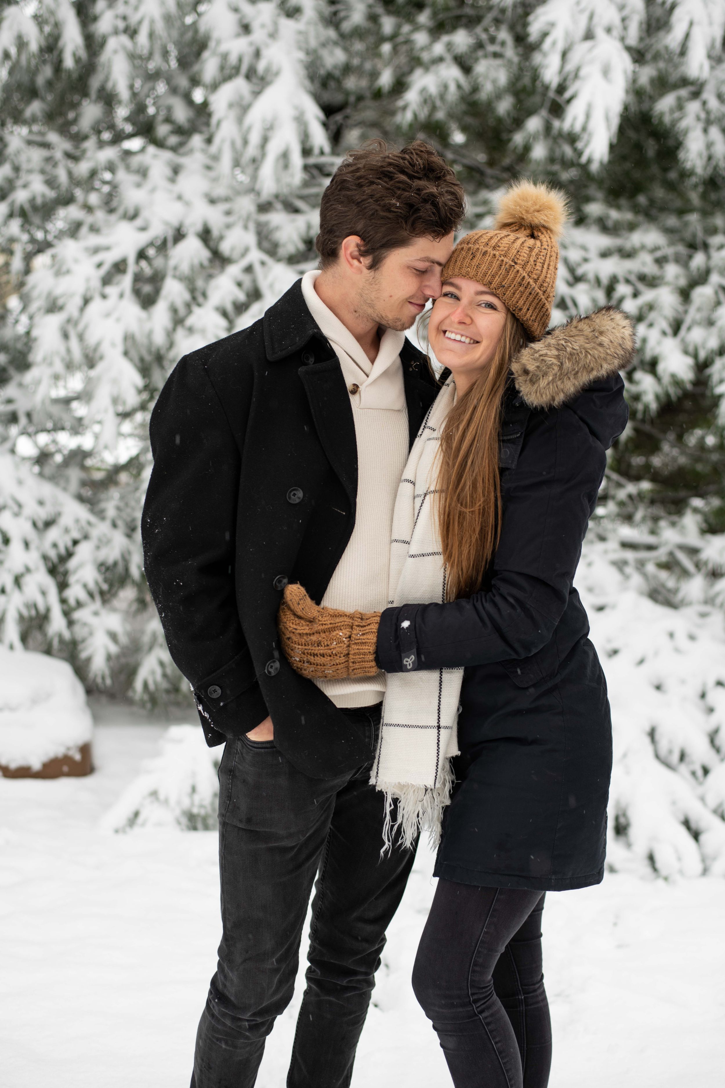 A couple in embracing and smiling in a snowy forest setting with snow-covered evergreen trees in background.