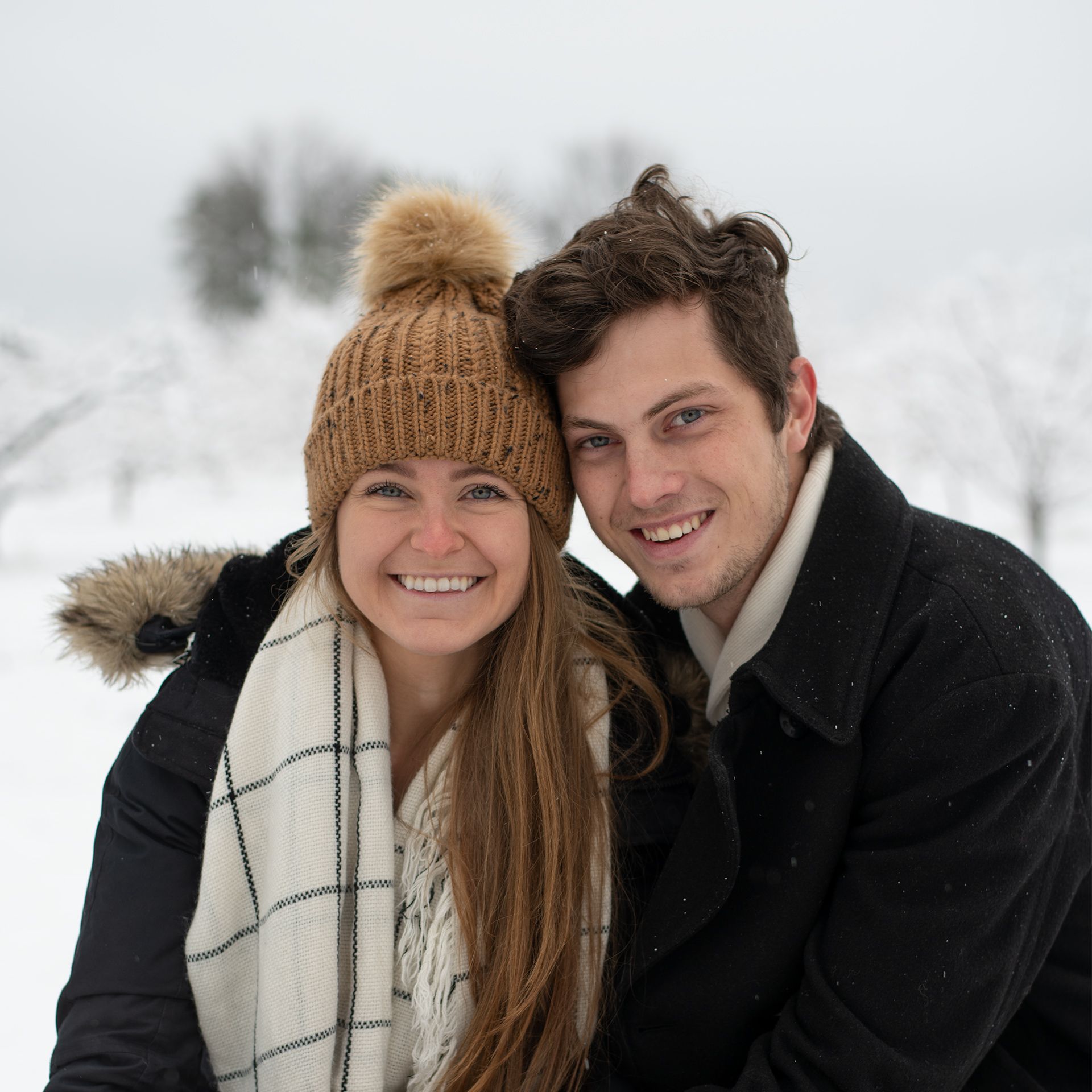 Two people smiling warmly while posing together in a snowy winter landscape.
