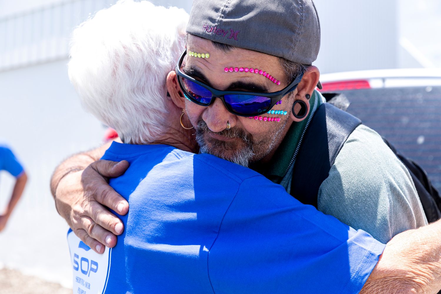 An emotional embrace between two people at an outdoor event, one wearing sunglasses and a cap.