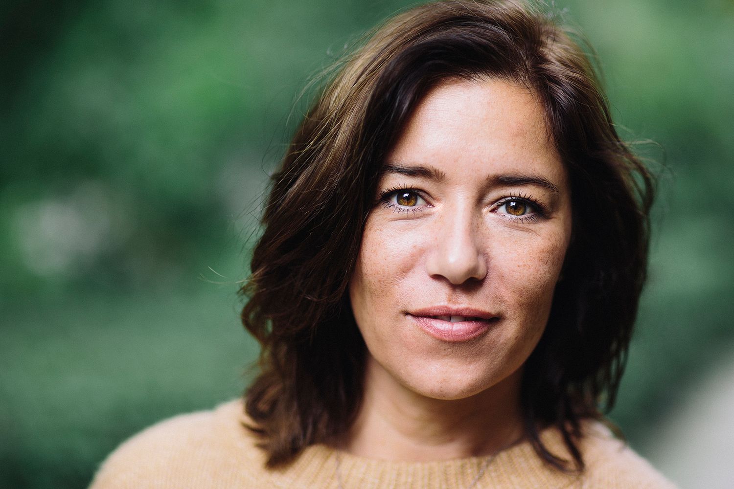 Portrait of a person with shoulder-length dark hair wearing a tan sweater against a blurred green background.