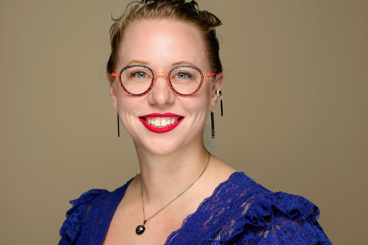 A portrait of someone wearing a blue top, round glasses and bright red lipstick against a neutral background.
