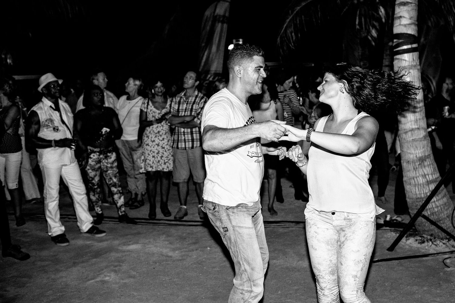 Black and white photo of a couple dancing at an outdoor gathering among palm trees.