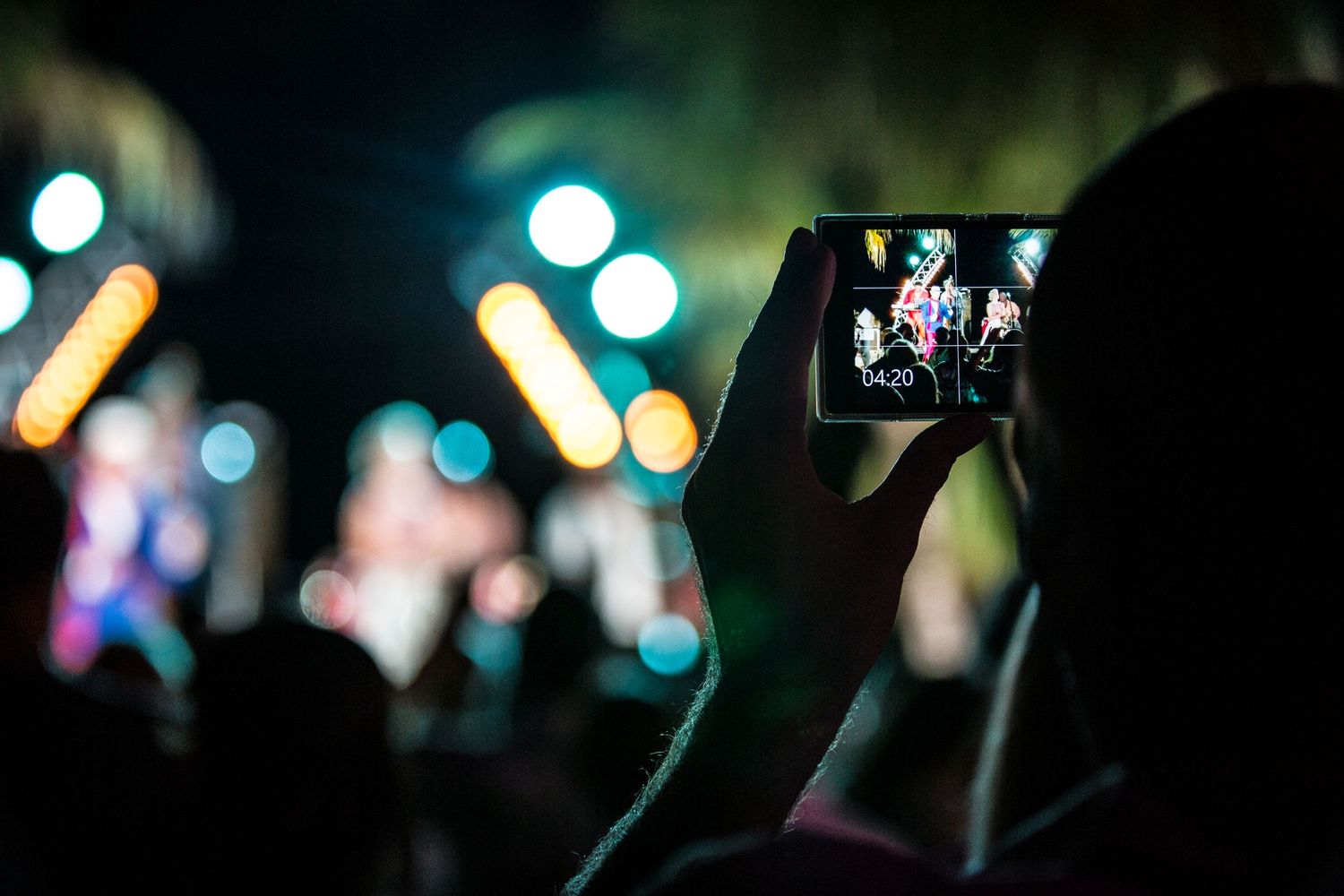 Hand holding up a smartphone to record a concert with blurred colorful bokeh lights in the background.