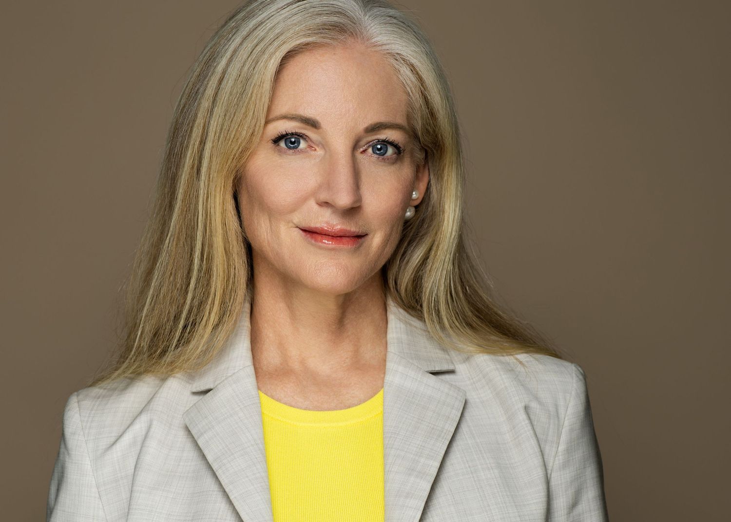 Professional portrait shot against brown background showing person with long blonde hair wearing grey blazer and yellow top.
