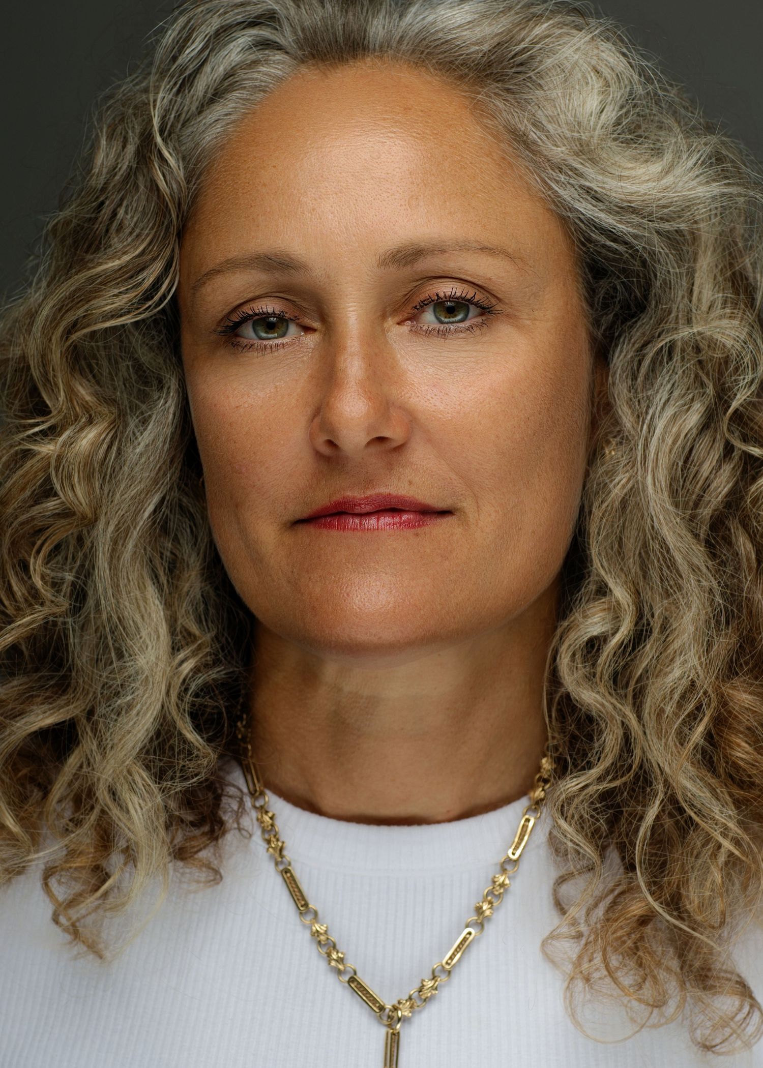 Woman with long curly gray hair wearing white top and gold necklace poses for portrait against dark background.