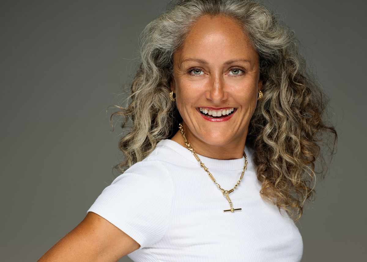 Smiling person wearing white t-shirt and gold necklace with long curly gray hair against gray background.