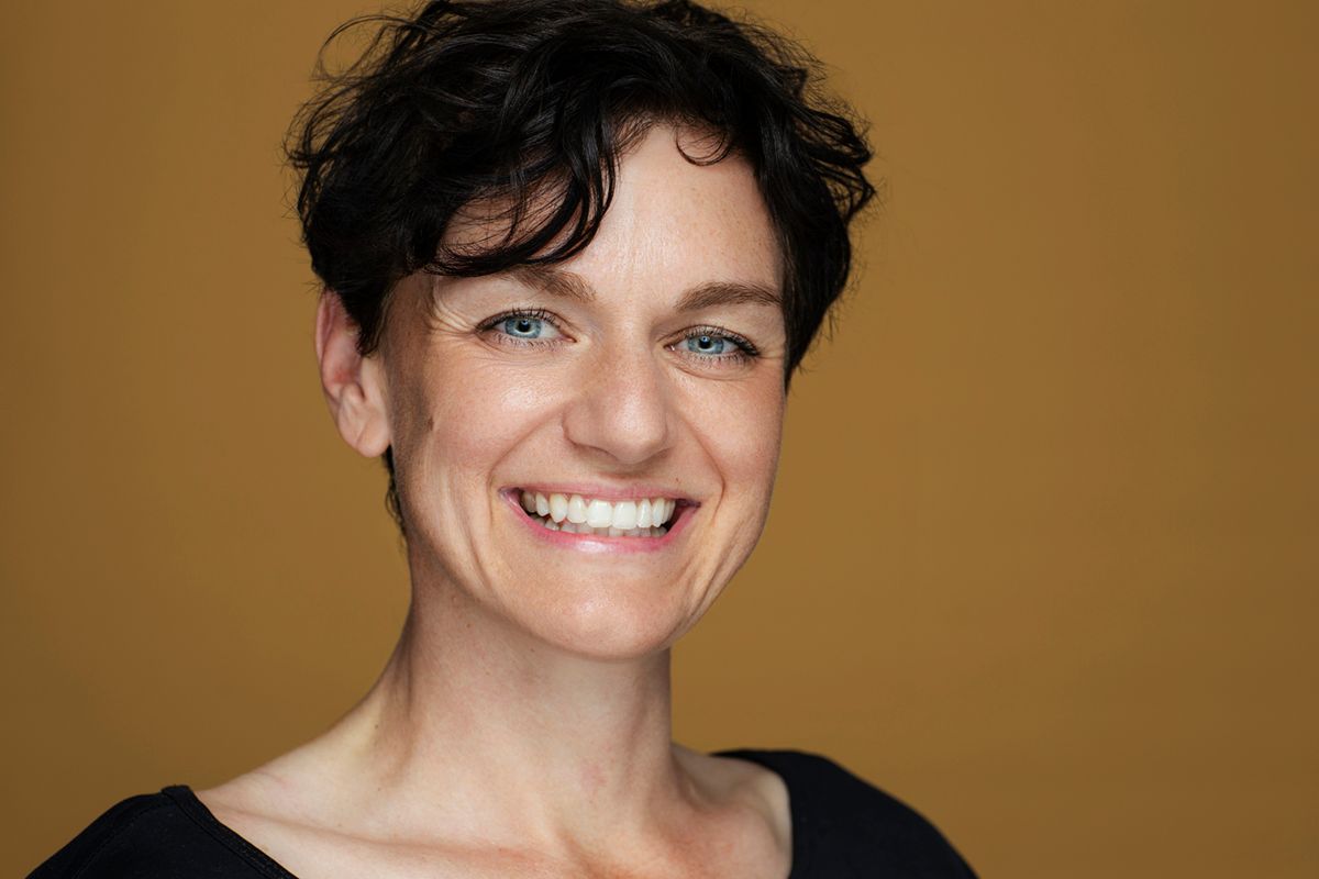 A close-up portrait shot of a person with dark curly hair and a warm smile against a brown background.