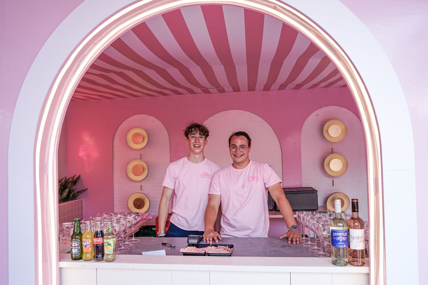 Two people in white chef coats stand behind a pink-themed bar counter with decorative plates on the wall.