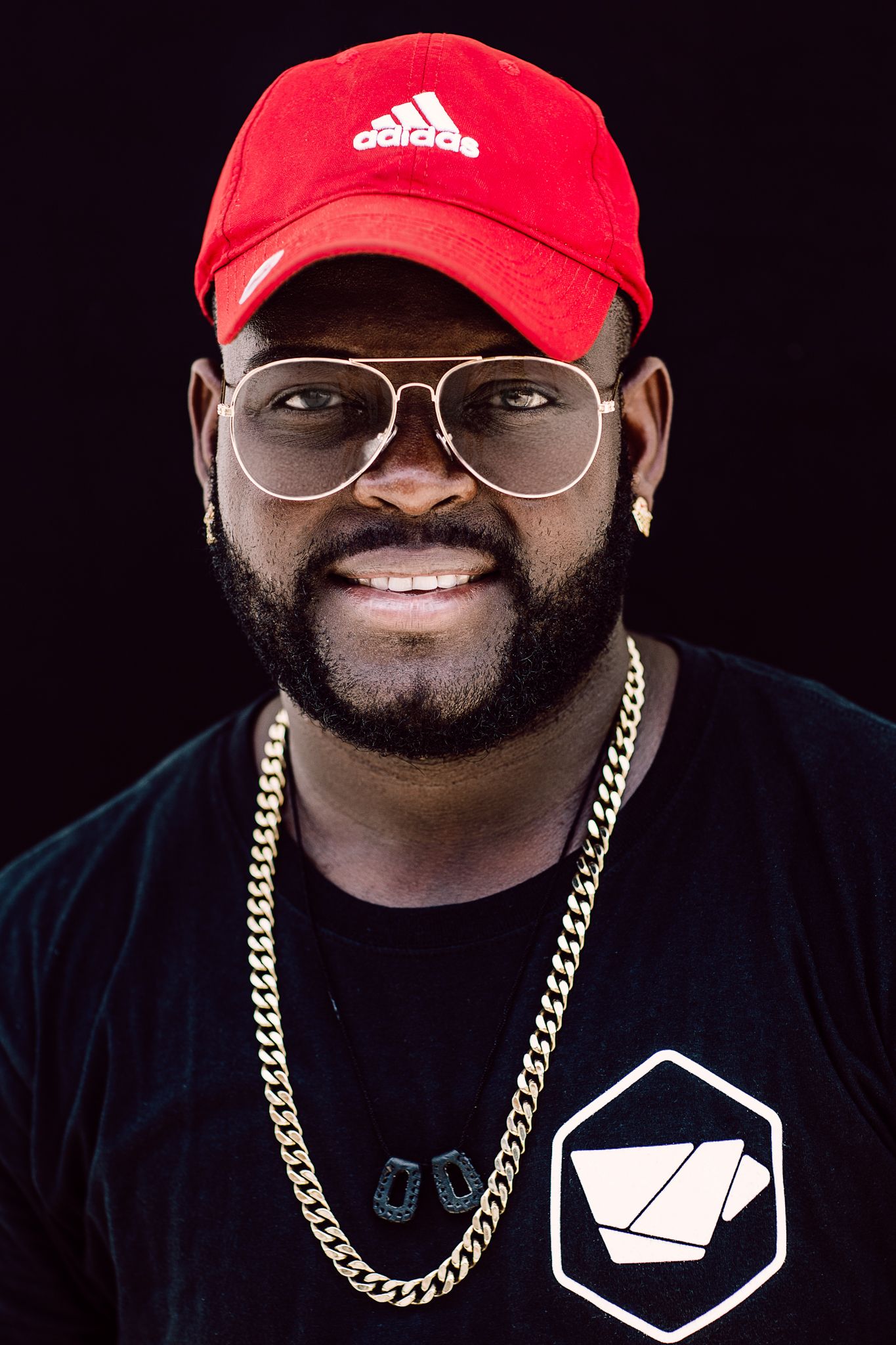 Portrait against dark background showing person wearing red Adidas cap, aviator glasses, gold chain and black t-shirt with geometric logo.