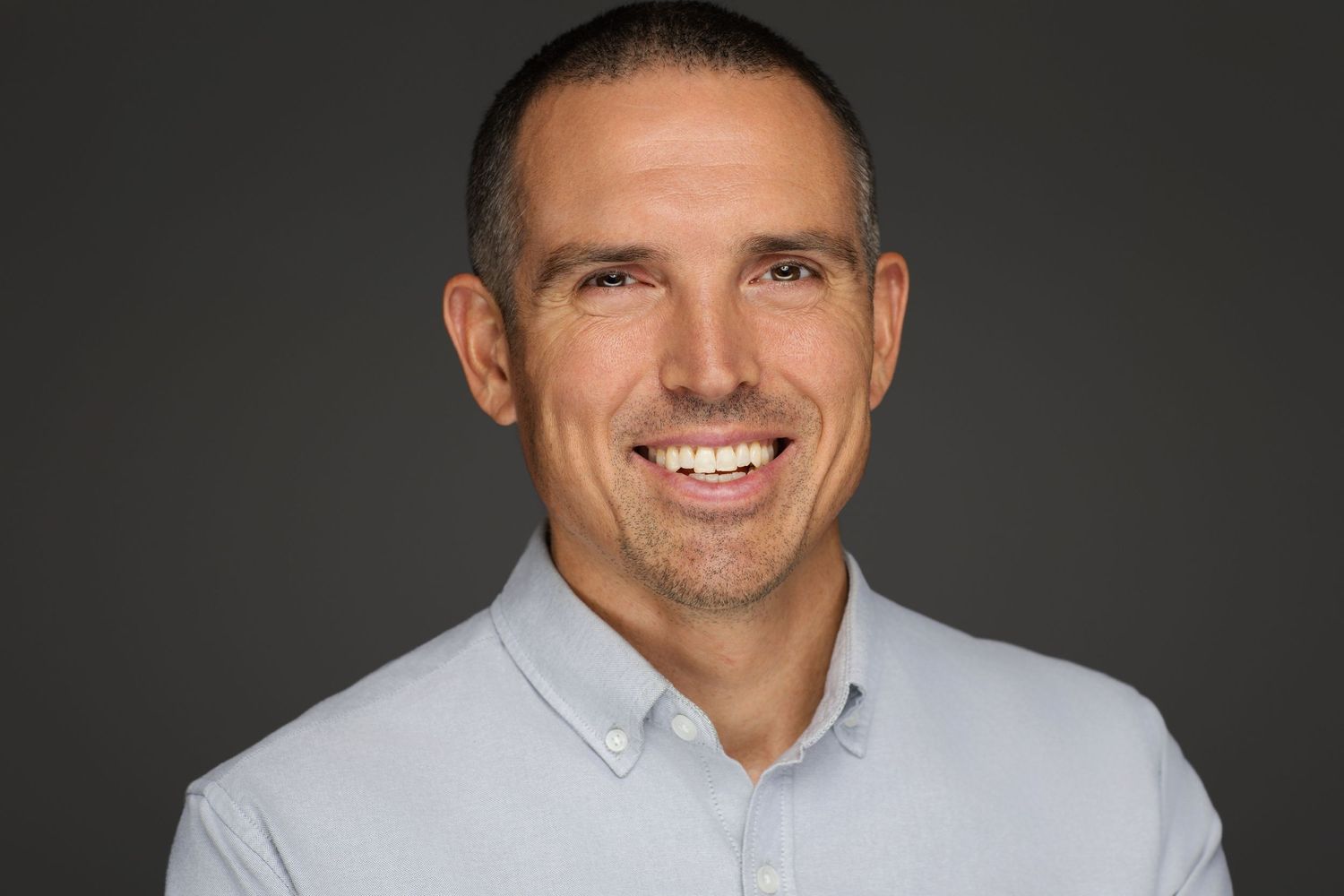A professional headshot of a person wearing a light gray collared shirt smiling warmly against a dark background.