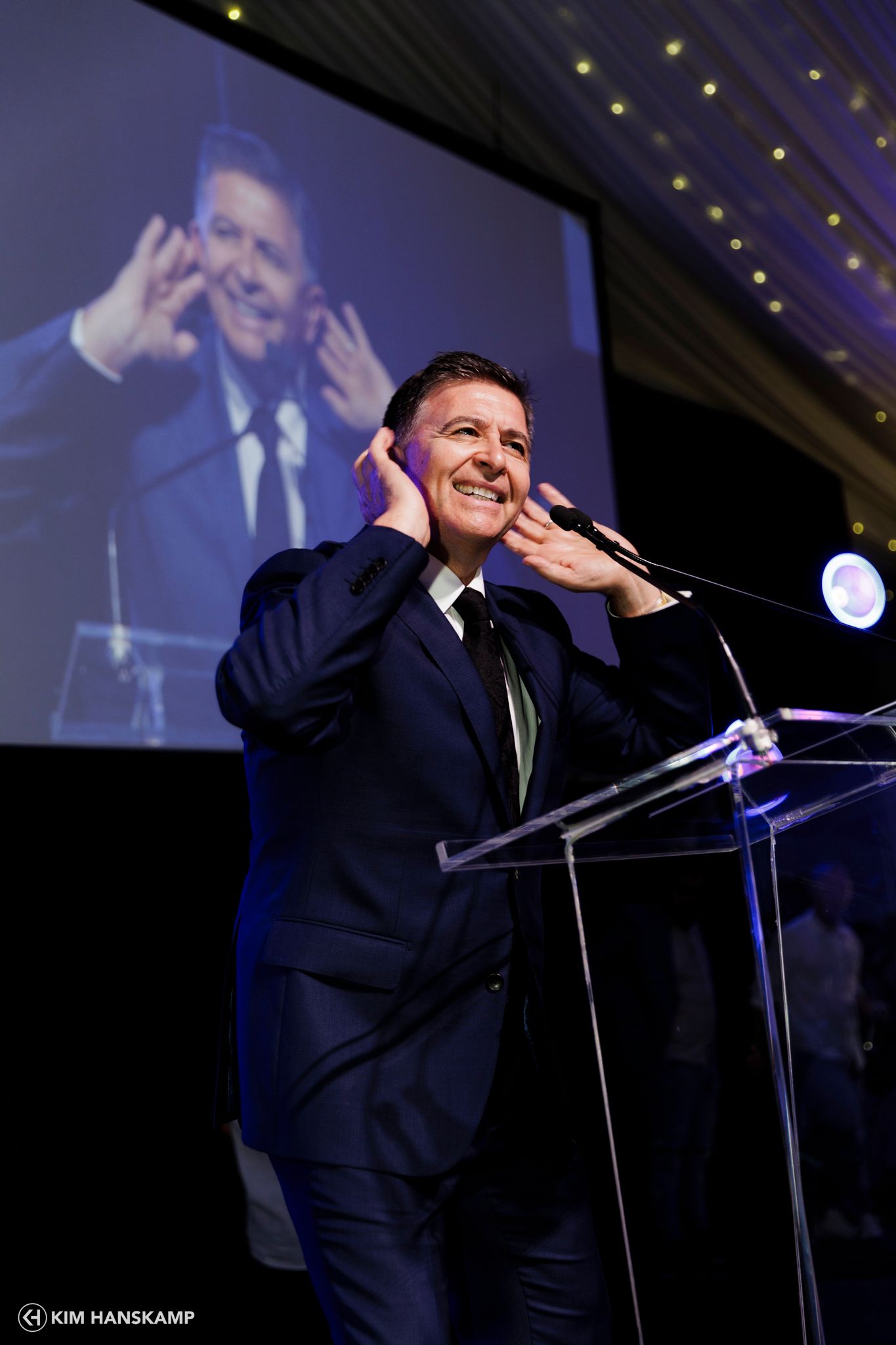 Speaker gesturing at podium during presentation with projection screen in background.